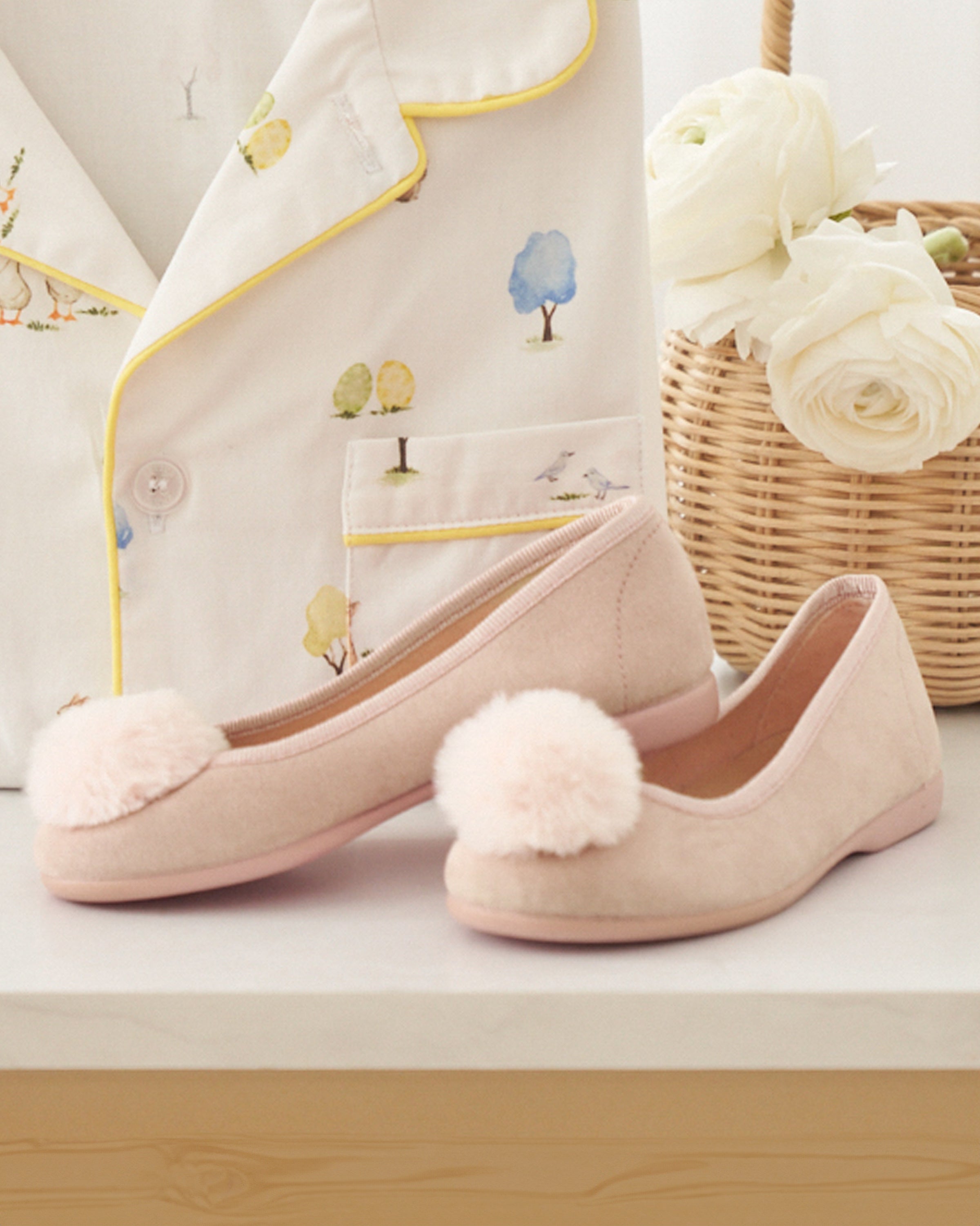 Petite Plumes Kids Juliette Slippers in Peach Suede with Festive Poms sit elegantly on a table next to a cream pajama top featuring small tree designs, while a wicker basket filled with white flowers adds charm in the background.