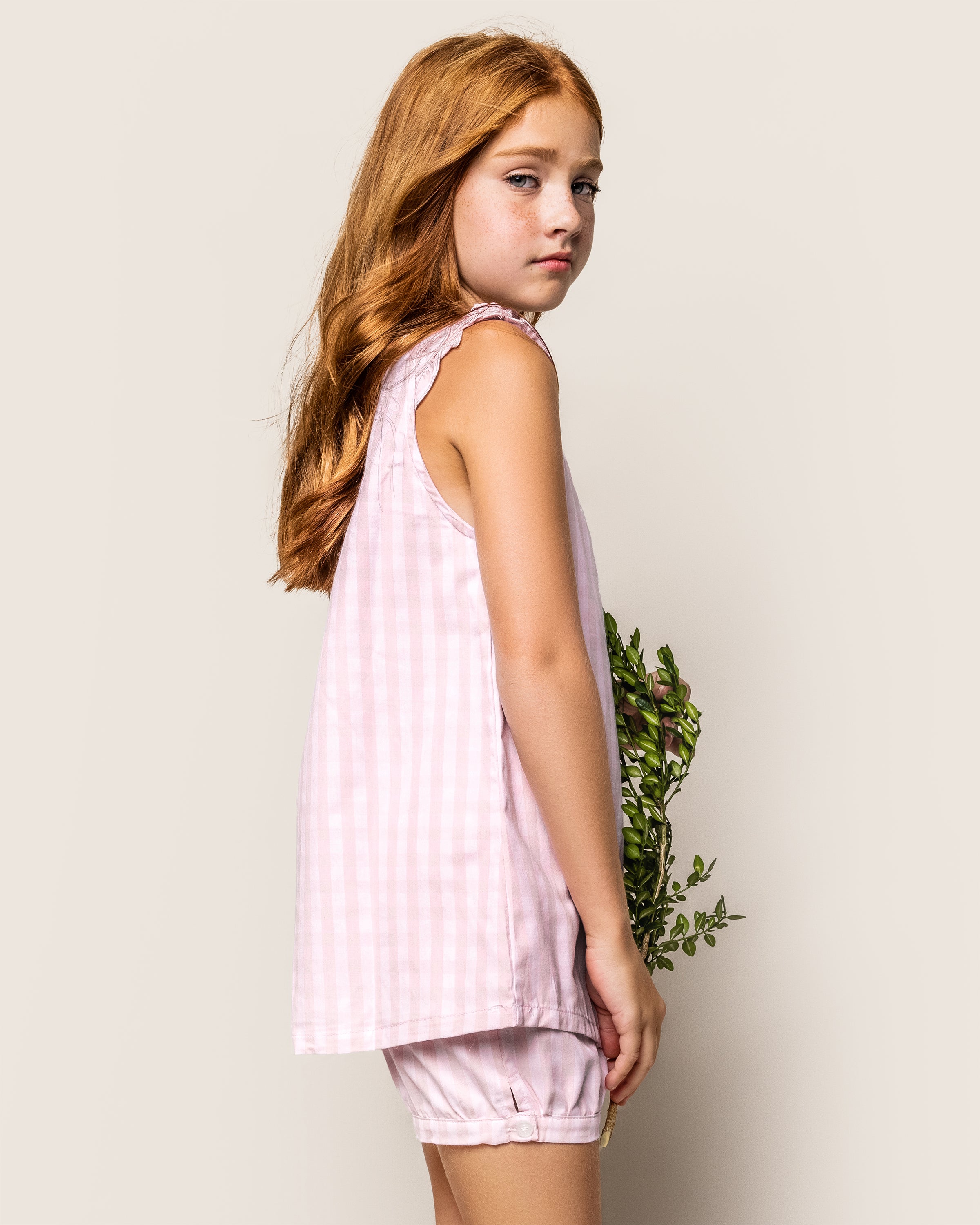 A young girl with long red hair stands in profile, wearing the Girls Twill Amelie Short Set in Pink Gingham by Petite Plume. Her stylish pink and white gingham outfit complements the small branch with green leaves she holds against a plain background.