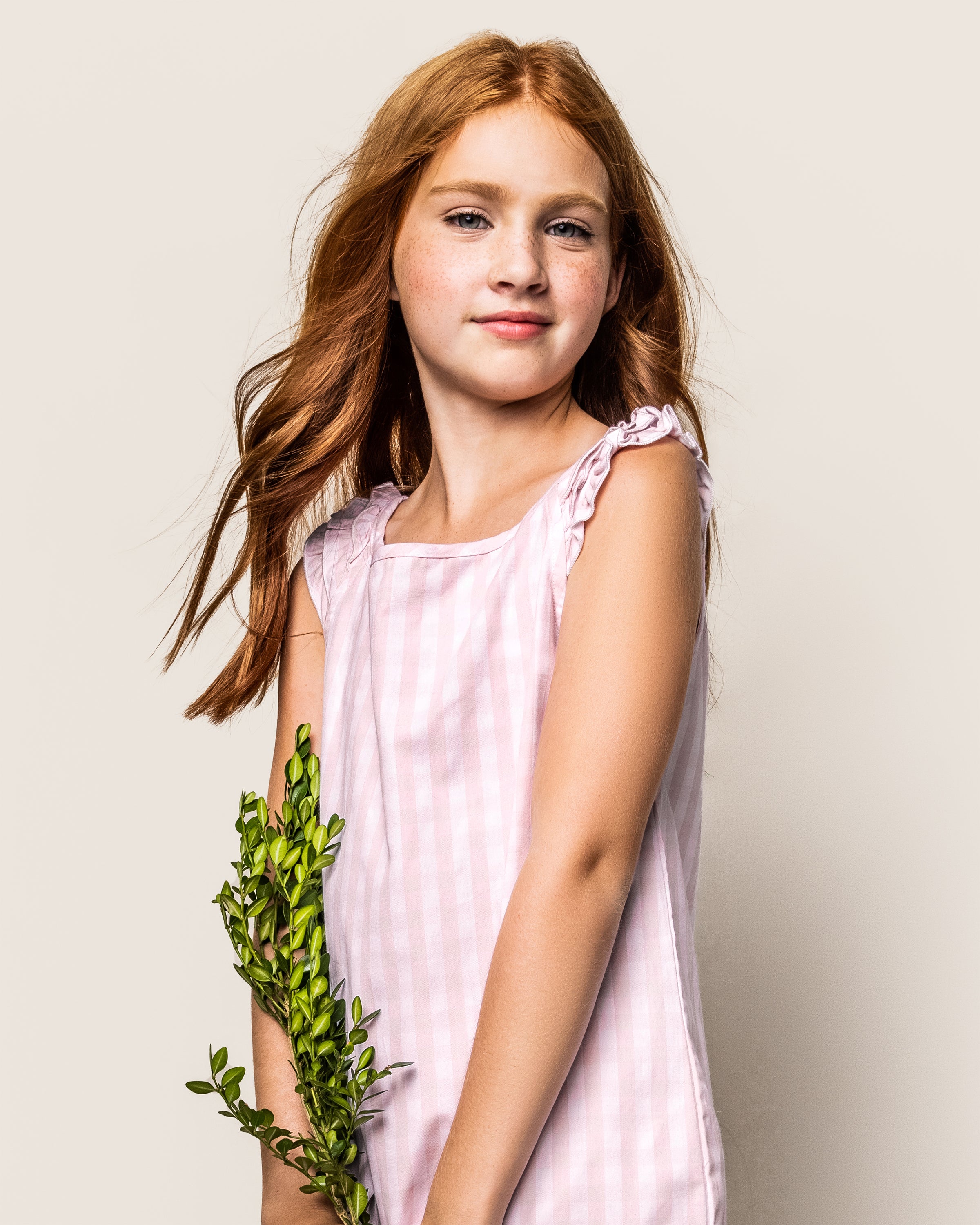 A young girl with long red hair wears the Petite Plume Girls Twill Amelie Short Set in pink gingham. Holding green foliage in her left hand against a plain background, her calm and confident expression epitomizes elegance reminiscent of luxurious sleepwear.