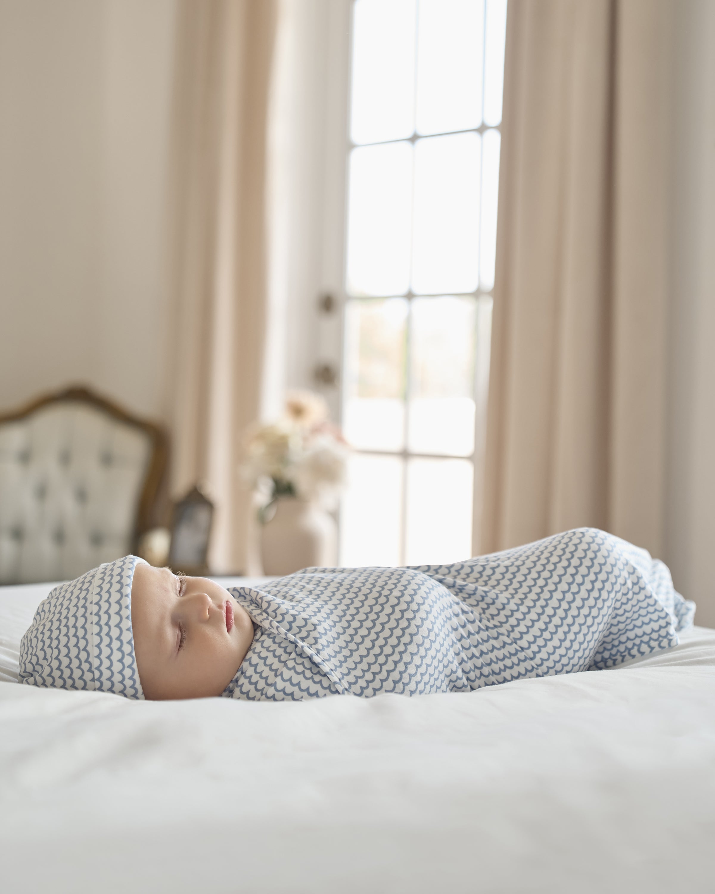 A baby sleeps peacefully on a bed, wrapped in Petite Plumes The Hospital Stay Luxe Set in Periwinkle & La Mer—featuring a matching cap. The serene scene includes soft lighting, a tufted chair, flowers, and tall curtains against a bright window.