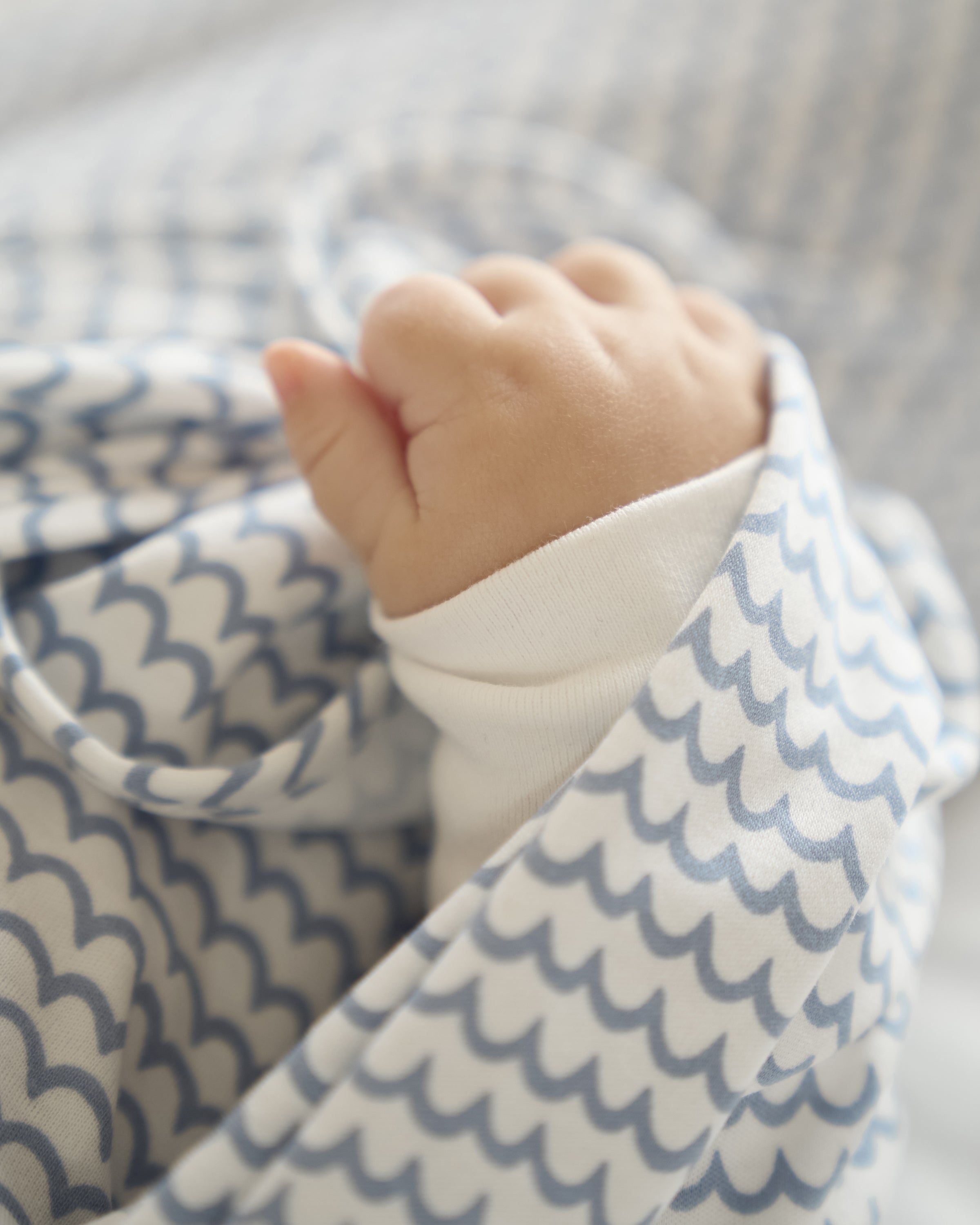 A babys hand peeks from a white sleeve, nestled in The Hospital Stay Luxe Set - Periwinkle & La Mer by Petite Plume, featuring a blanket with blue and white scallop designs, perfect for an exquisite gift presentation.