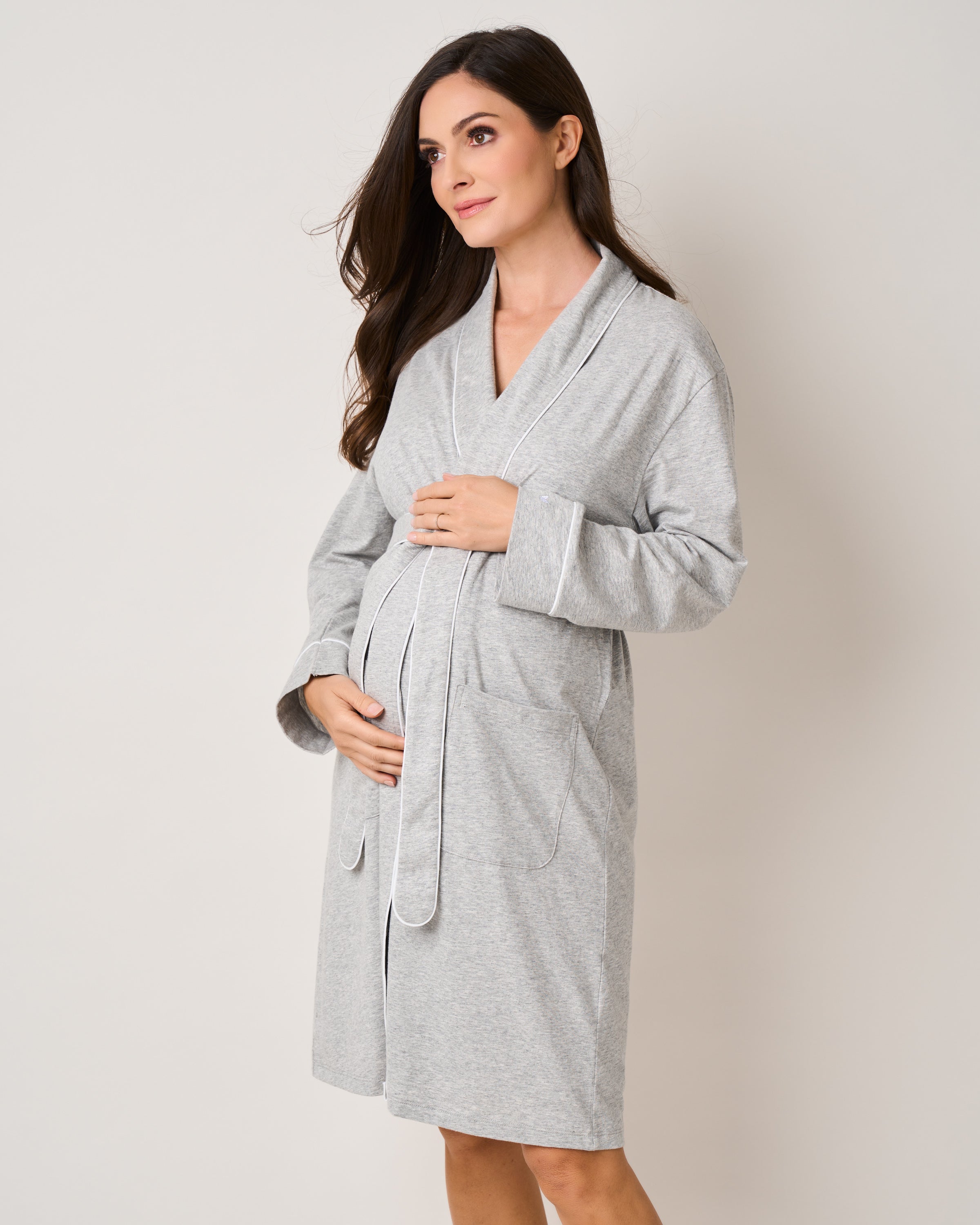 A pregnant woman, wrapped in The Cozy Maternity Set in Light Heather Grey by Petite Plume, cradles her belly. Her long brown hair cascades as she gazes sideways, embodying luxurious relaxation against the plain off-white backdrop—a perfect gift presentation.