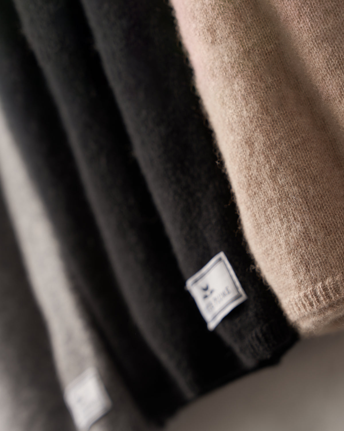 Close-up of folded sweaters in neutral tones: beige, black, gray, and taupe. Sweaters arranged diagonally with visible Petite Plume labels. The luxurious texture of the Womens Cashmere Wrap in Beige exudes comfort with its soft knit fabric.