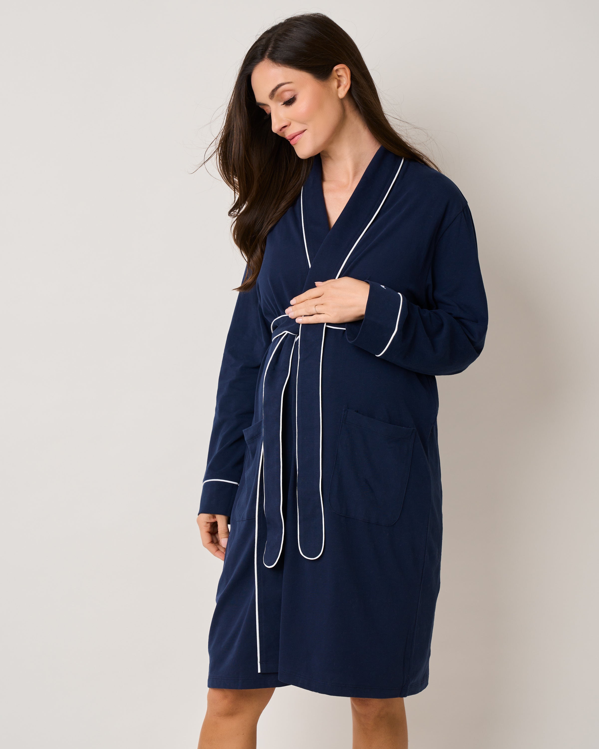 A woman stands against a plain background, wrapped in Petite Plumes The Cozy Maternity Set in Navy. Her long dark hair frames a gentle smile as she rests one hand on her chest. The set includes a navy robe with white piping, featuring a belt at the waist and two pockets.