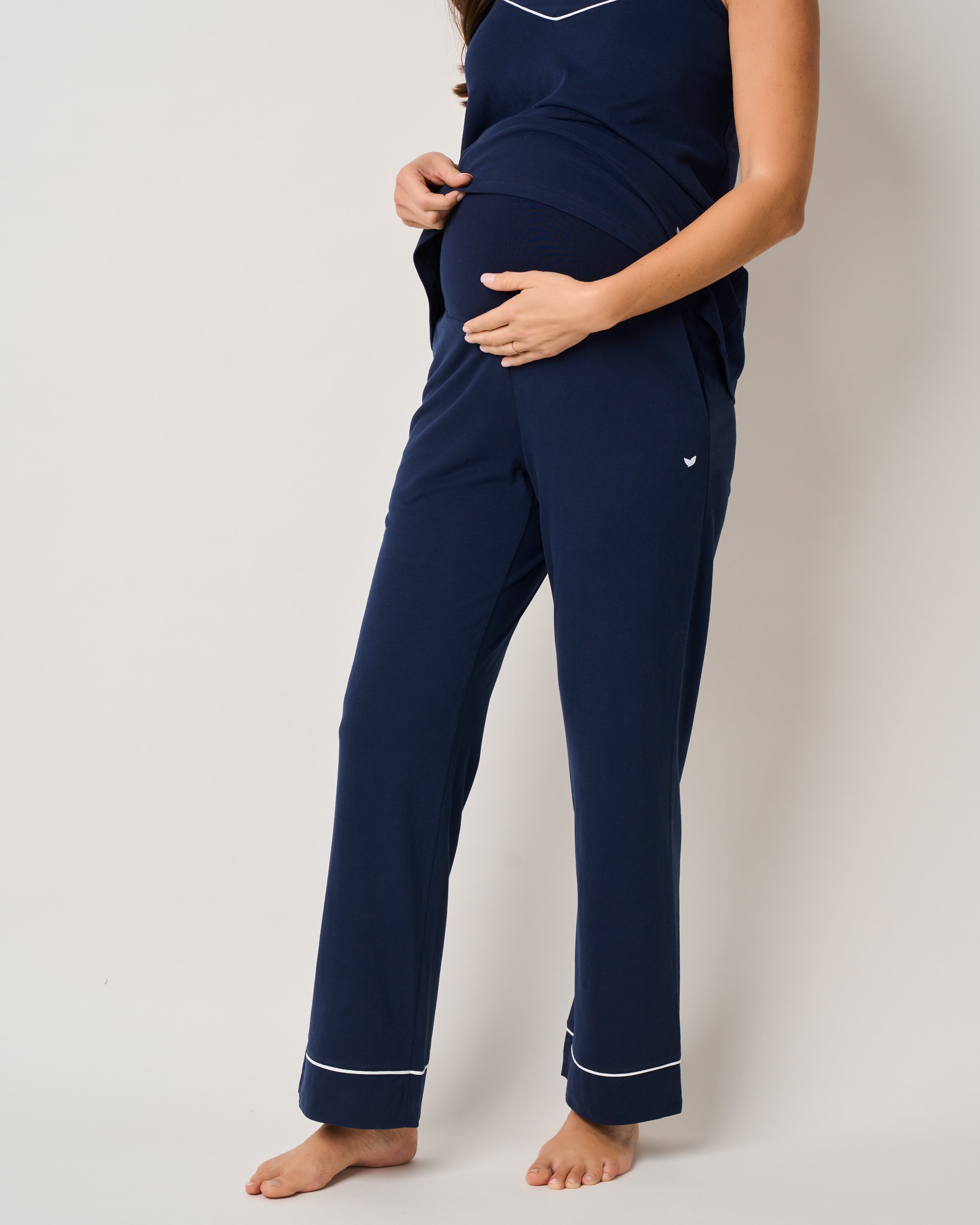 A pregnant woman in Petite Plumes The Basics Maternity Set in Navy with white trim gently holds her belly, barefoot against a plain white background, embodying the comfort and elegance of essential maternity wear.