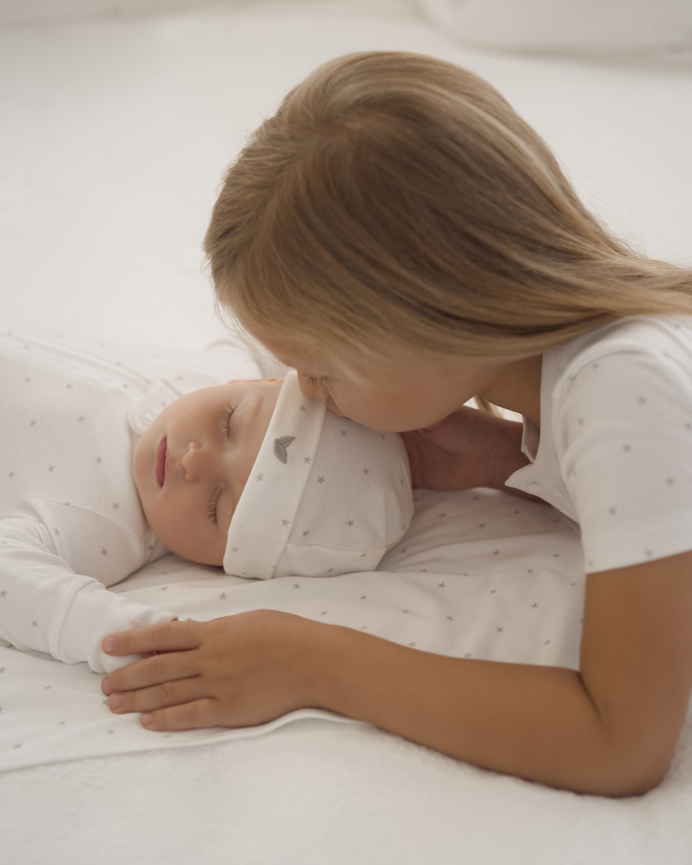 A young girl gently kisses a sleeping baby who rests on a white surface in Petite Plumes soft baby clothing made from Peruvian Pima cotton. The little one wears the Babys Pima Hat in Grey Stars, while the girl holds the hat and wears a white shirt.