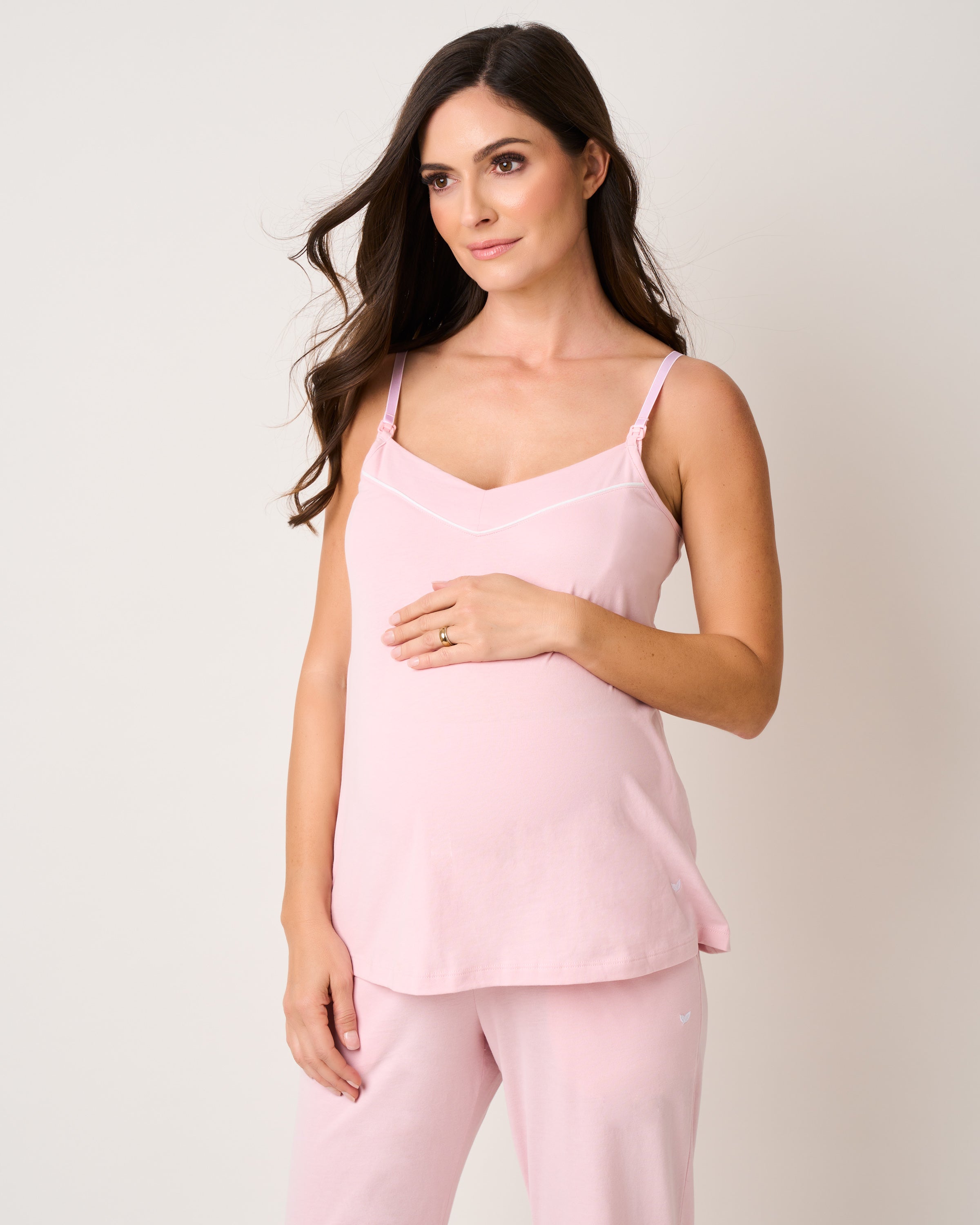A woman, possibly on her pregnancy journey, dons a Petite Plume Womens Pima Maternity Camisole in Pink while gently cradling her belly. Her long brown hair frames a slight smile as she stands against a plain beige background.