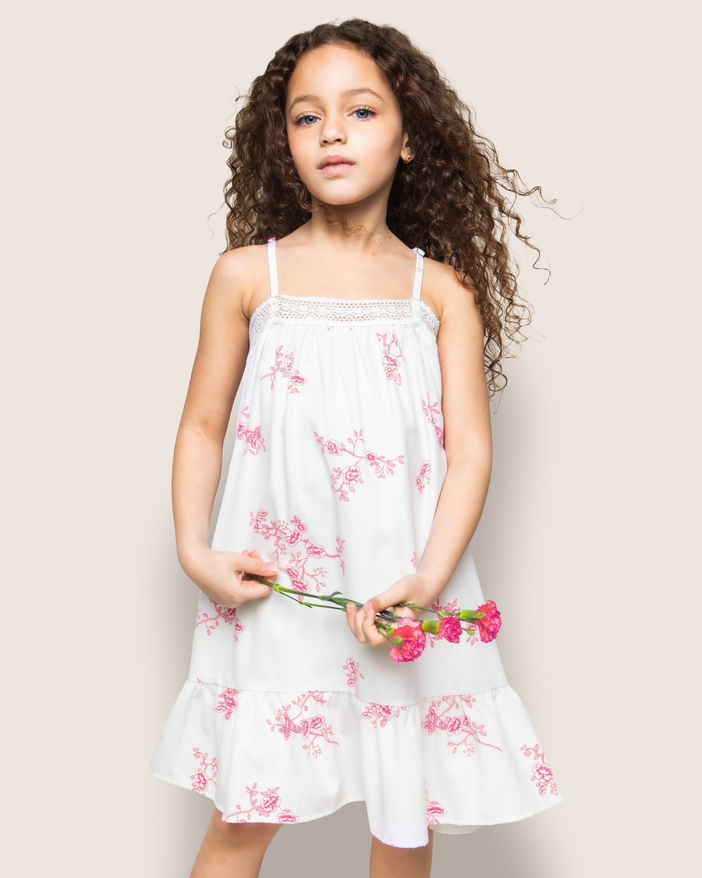 A young curly-haired girl models the Petite Plume Girls Twill Lily Nightgown in English Rose Floral. She holds pink flowers against a simple backdrop, showcasing the elegant design.