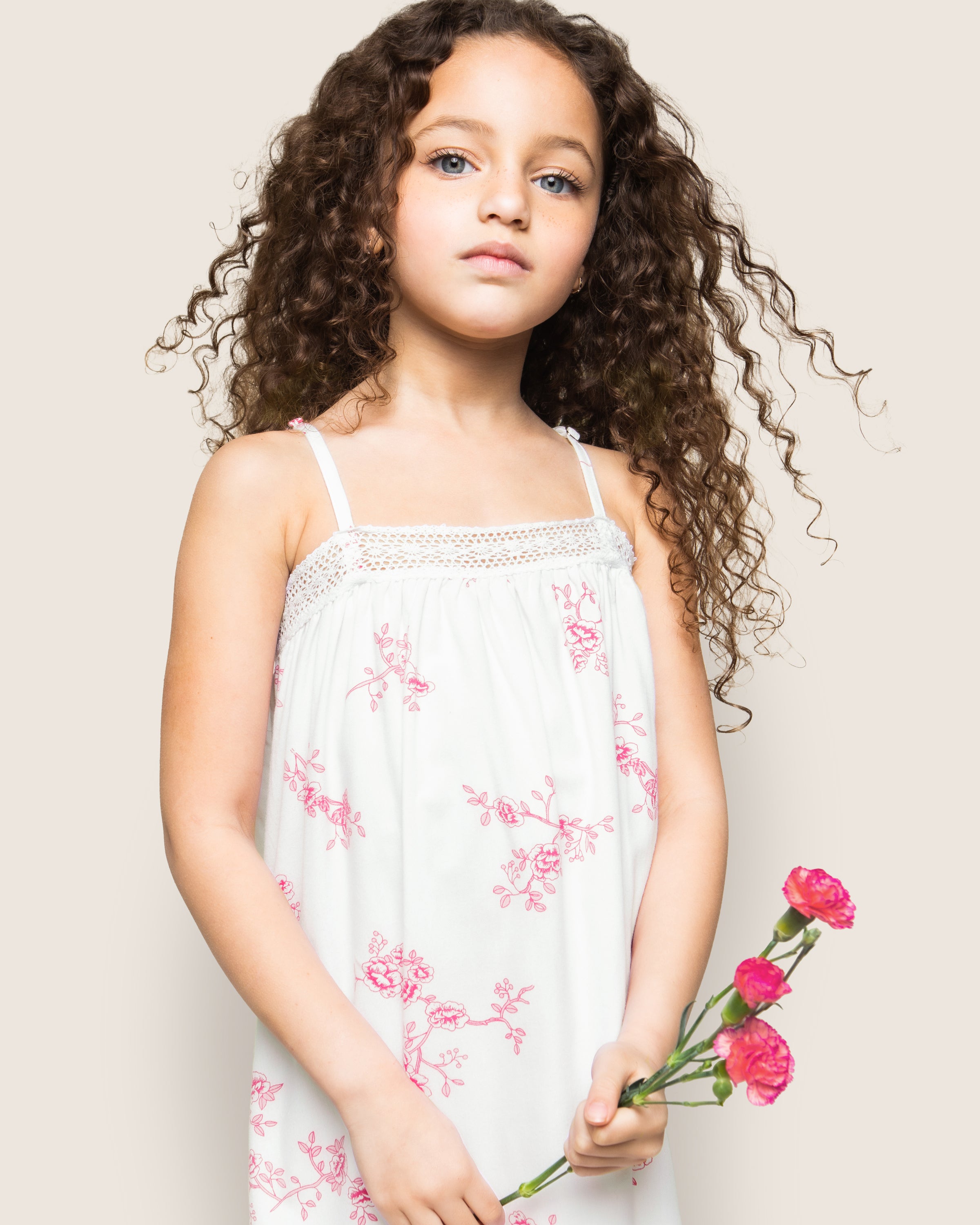 A young girl with curly hair wears Petite Plumes Girls Twill Lily Nightgown in English Rose Floral. She gracefully holds a small bouquet of red flowers against a plain, light-colored background.