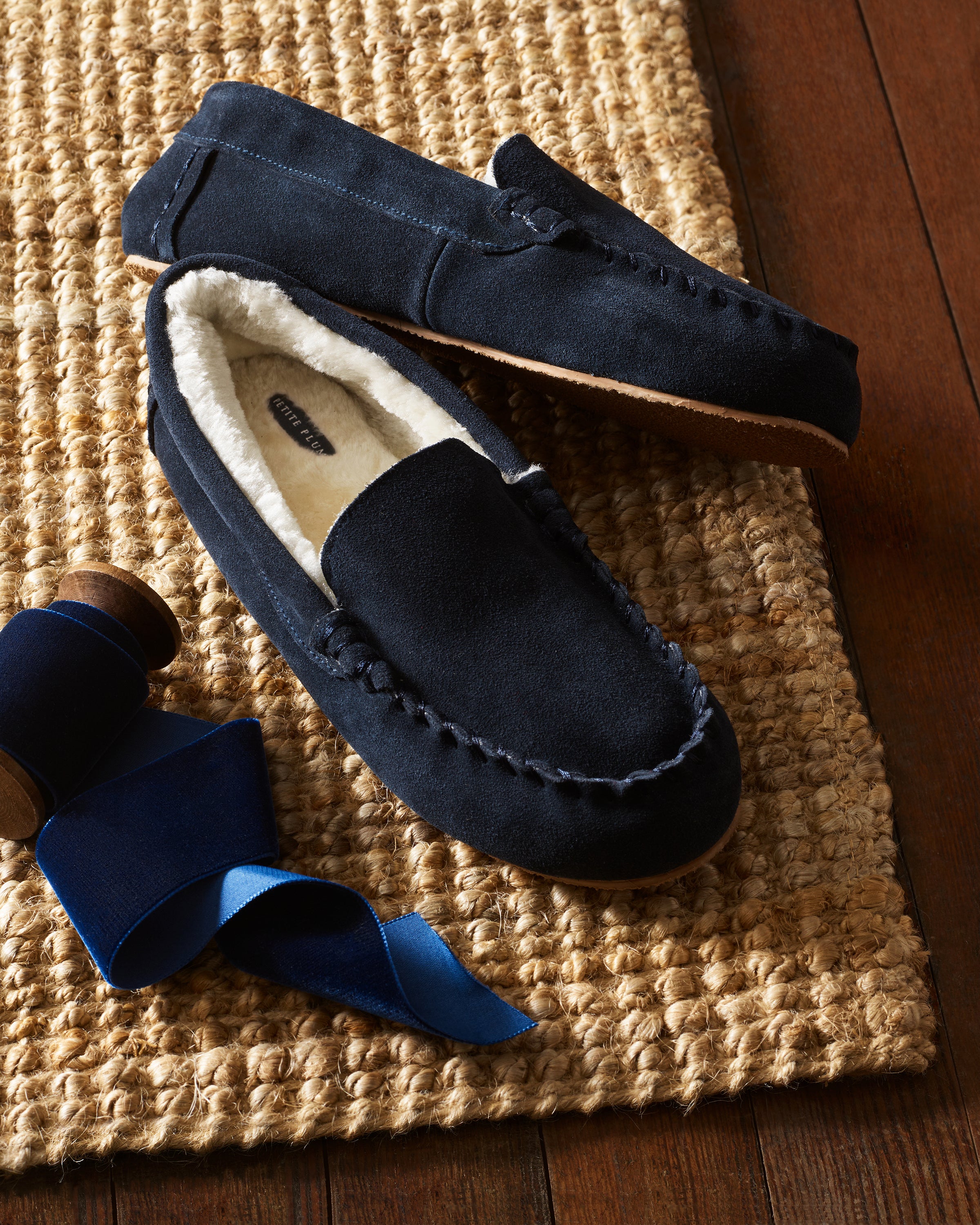 Petite Plumes Kid’s Suede Slippers in Navy, featuring fleece lining, rest on a woven rug beside an unrolled spool of royal blue ribbon. Set on a wooden floor, these ultra-soft moccasin slippers are showcased beautifully.