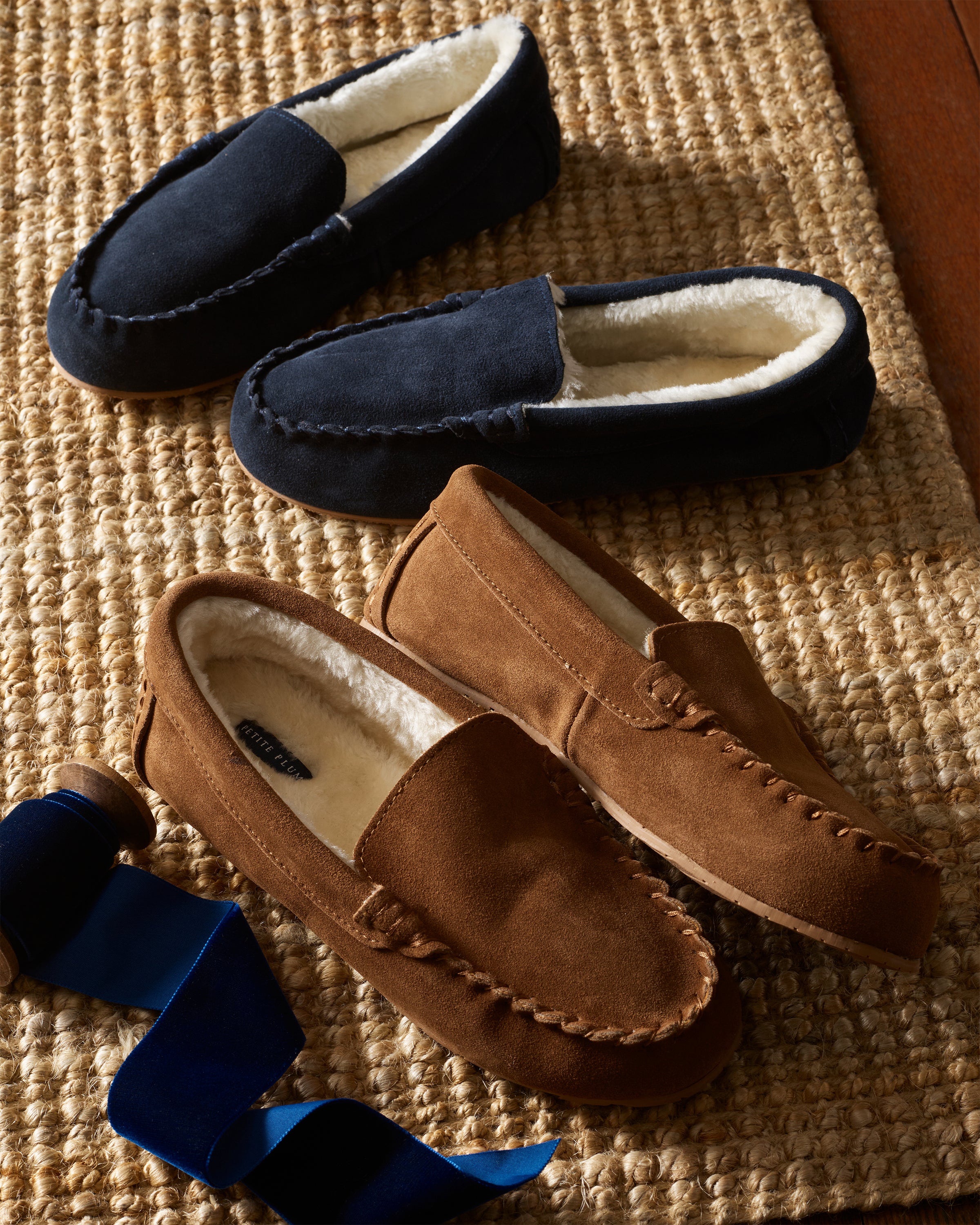 Two pairs of ultra-soft moccasin slippers, one Petite Plume Kids Suede Slippers in Navy and the other a brown version, rest on a woven rug. Both are lined with cream-colored faux fur fleece. A spool of dark blue ribbon lies nearby on the rug.