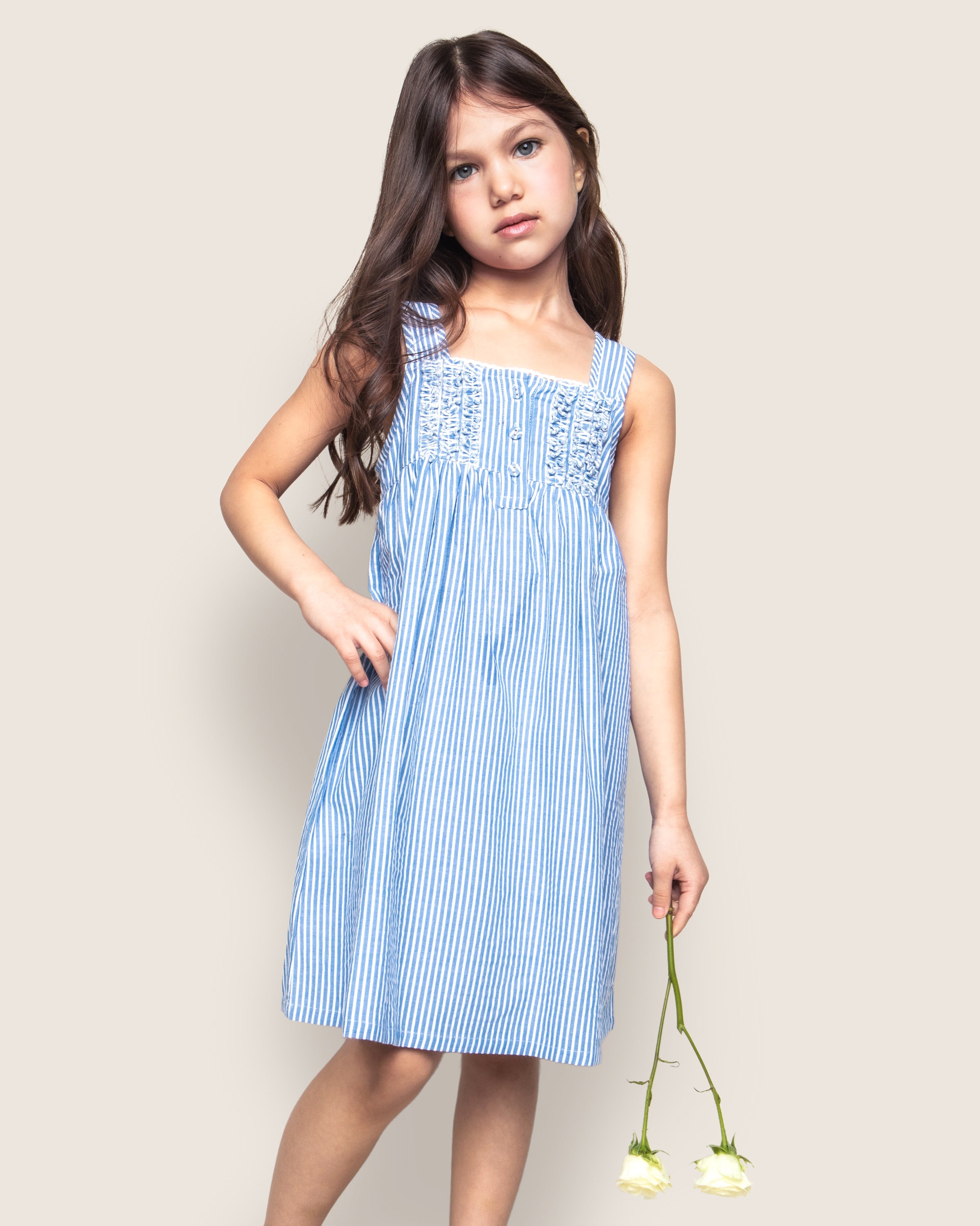 A young girl with long dark hair, wearing Petite Plumes Girls Twill Charlotte Nightgown in French Blue Seersucker, holds two white roses. She stands against a neutral background, gazing at the camera with a gentle expression.