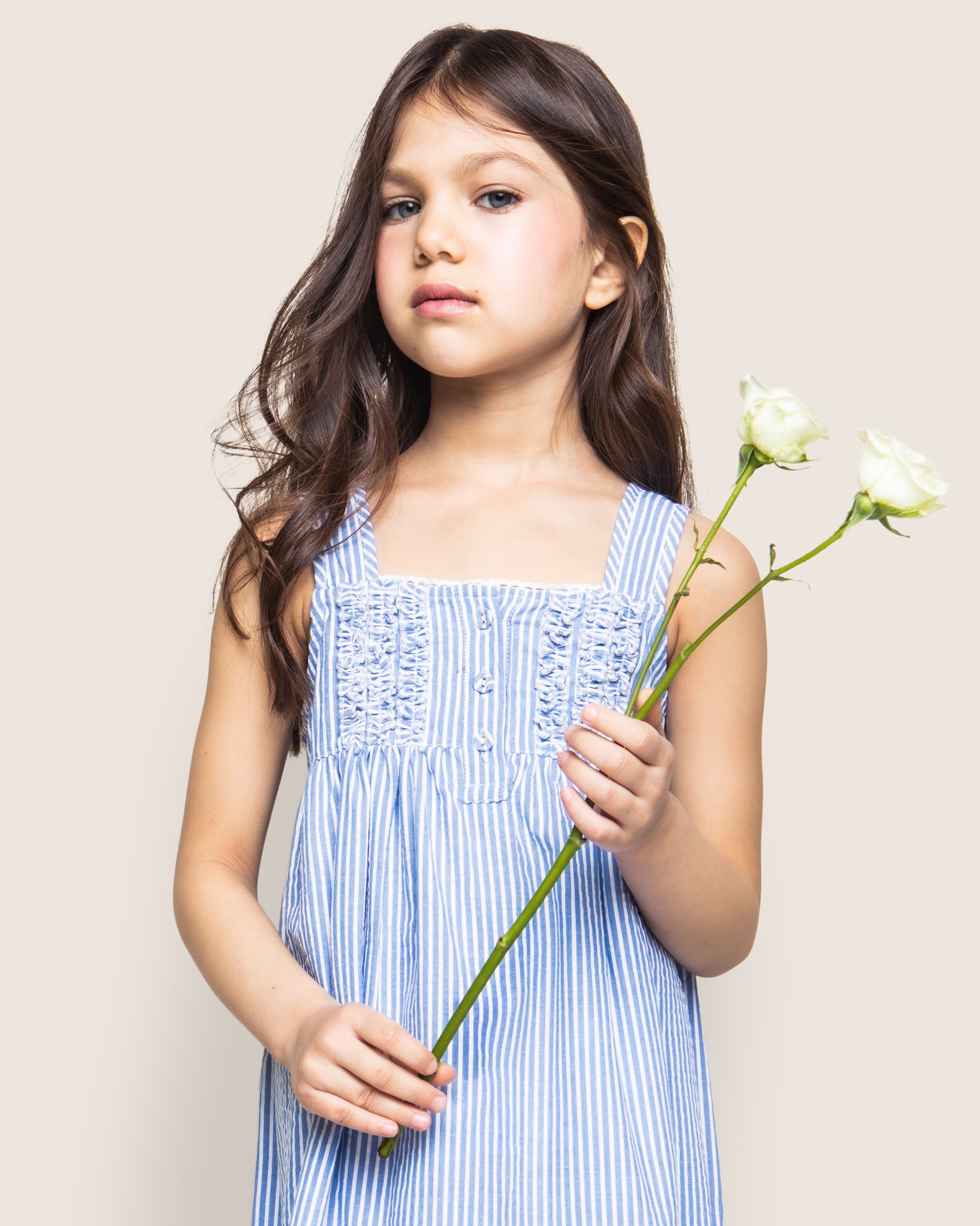 A young girl with long brown hair wears the Petite Plume Girls Twill Charlotte Nightgown in French Blue Seersucker and holds two white flowers, standing against a plain background and looking slightly to the side.