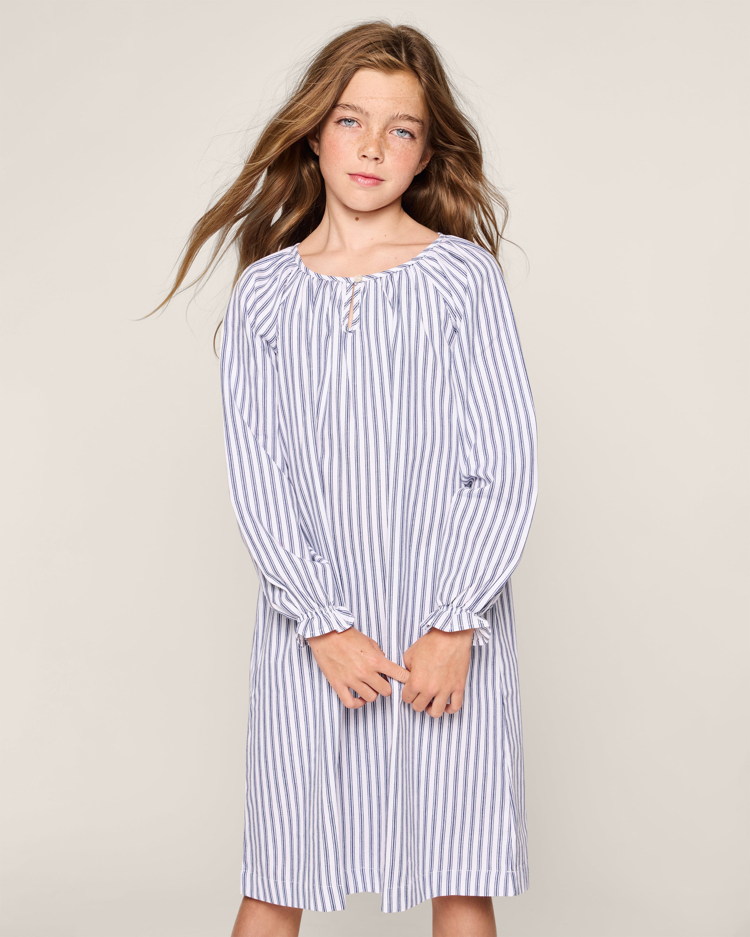 A young girl with long brown hair wearing Petite Plumes Girls Twill Delphine Nightgown in Navy French Ticking, crafted from flame-retardant fabric, stands against a plain background with a relaxed expression, facing forward.