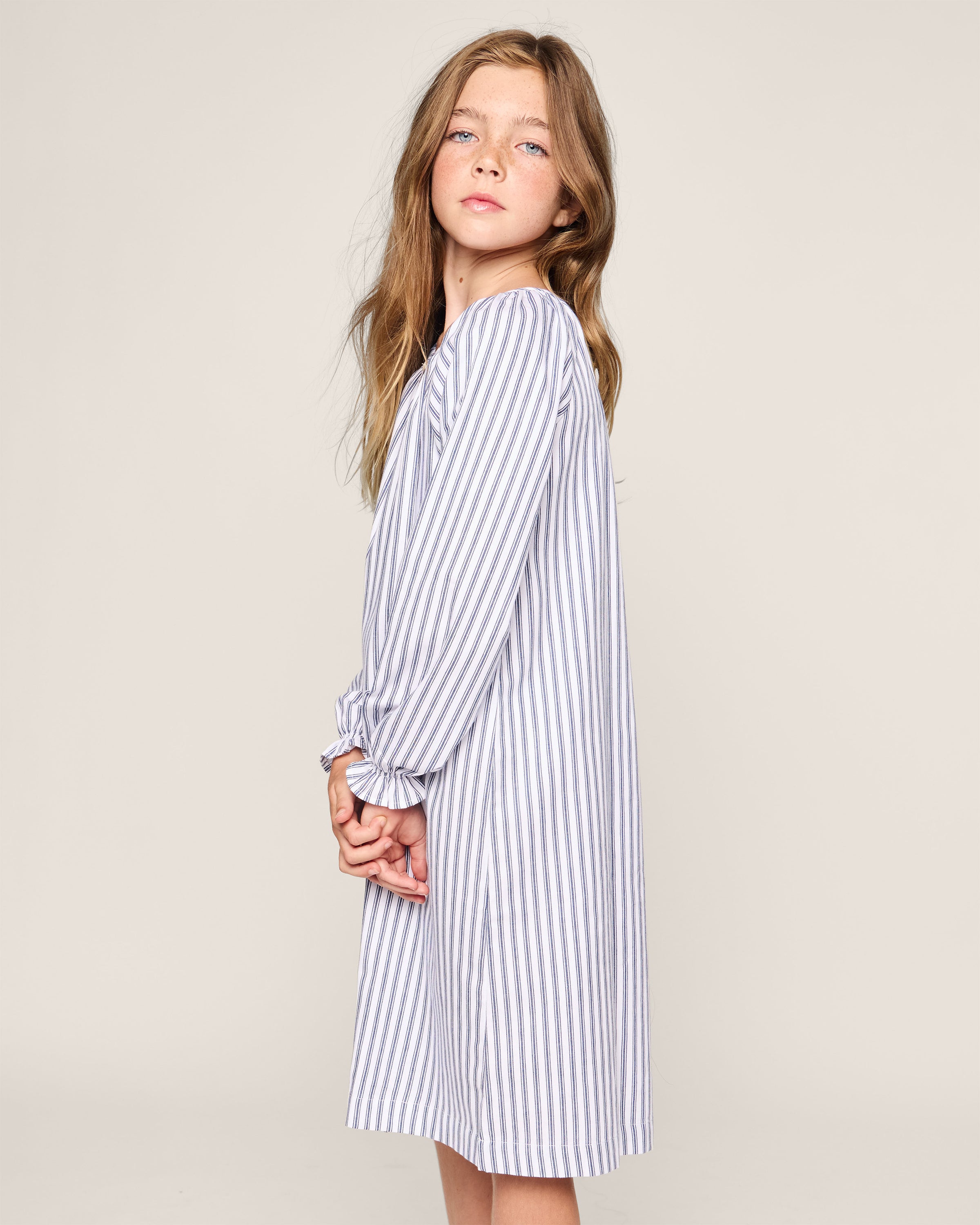 A young girl stands in profile, wearing a Petite Plume Girls Twill Delphine Nightgown in Navy French Ticking. Her long hair cascades down with hands clasped behind her back. The plain background highlights the elegance and safety of this childrens sleepwear made from flame-retardant fabric.