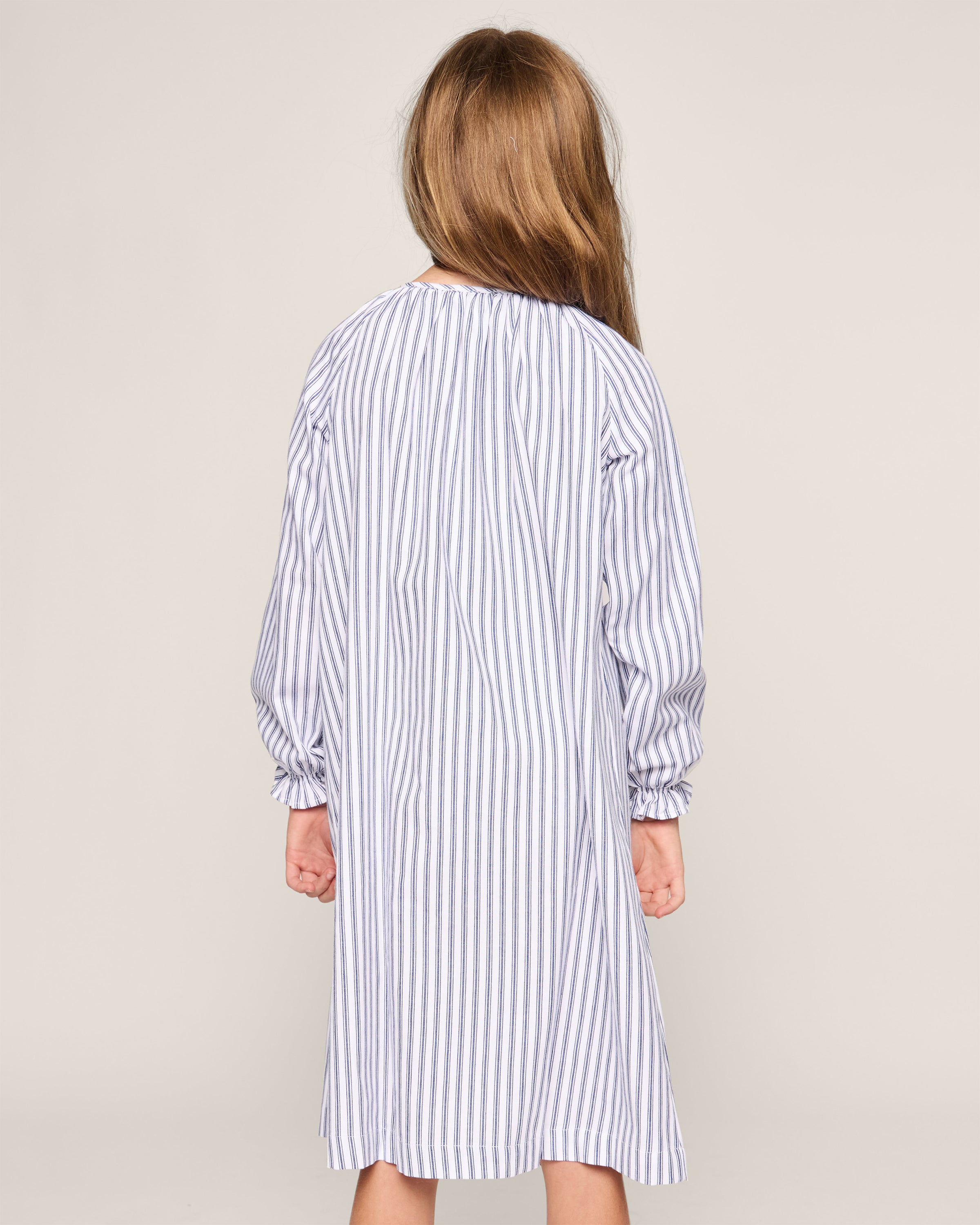A young girl with long hair facing away wears Petite Plumes Girls Twill Delphine Nightgown in Navy French Ticking, made from flame-retardant fabric. The sleepwear features vertical blue and white stripes against a plain, light-colored background.