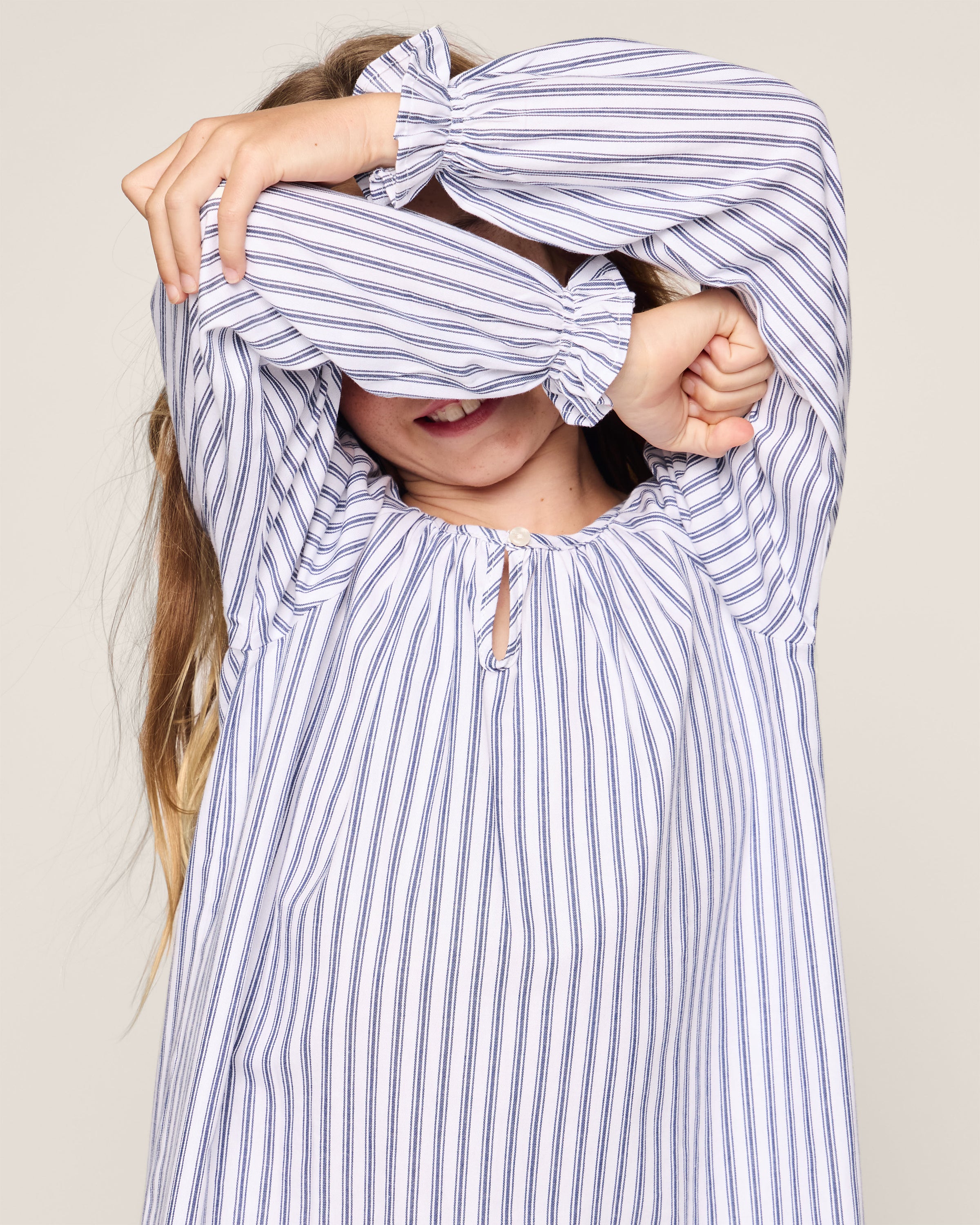 A child playfully covers their face with their hands, wearing Petite Plumes Girls Twill Delphine Nightgown in Navy French Ticking. Only the mouth and chin peek out against a plain light background.