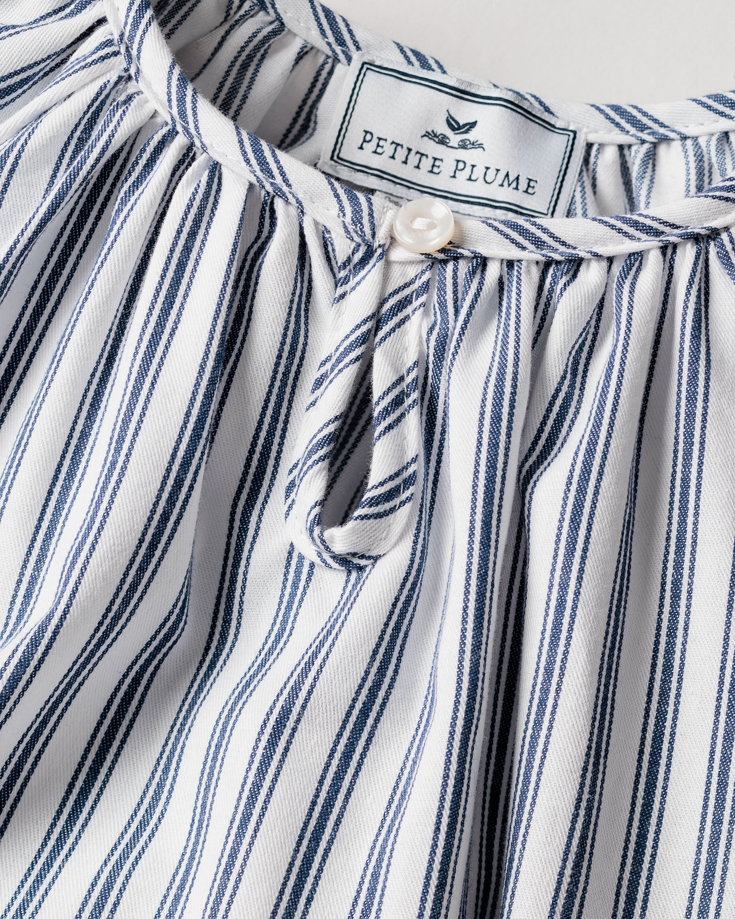 The Girls Twill Delphine Nightgown in Navy French Ticking by Petite Plume features blue and white vertical stripes. It is gathered at the top with a button loop closure and crafted from flame-retardant material, ensuring safety in childrens sleepwear. The brand label is visible at the neck area.