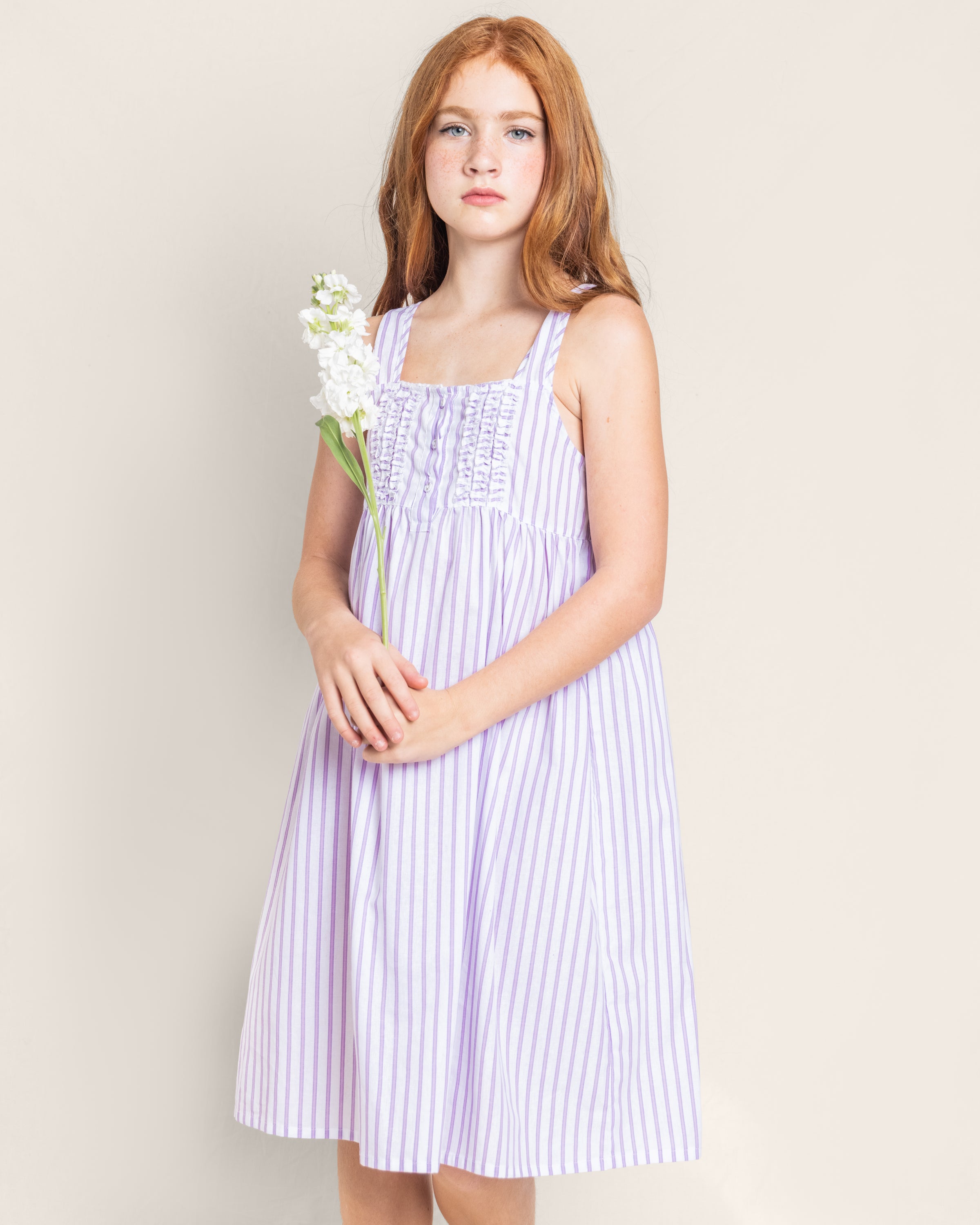 A girl with long red hair in a sleeveless Petite Plume Girls Twill Charlotte Nightgown in Lavender French Ticking holds white flowers. She stands against a plain beige background, exuding elegance and simplicity as she gazes forward.