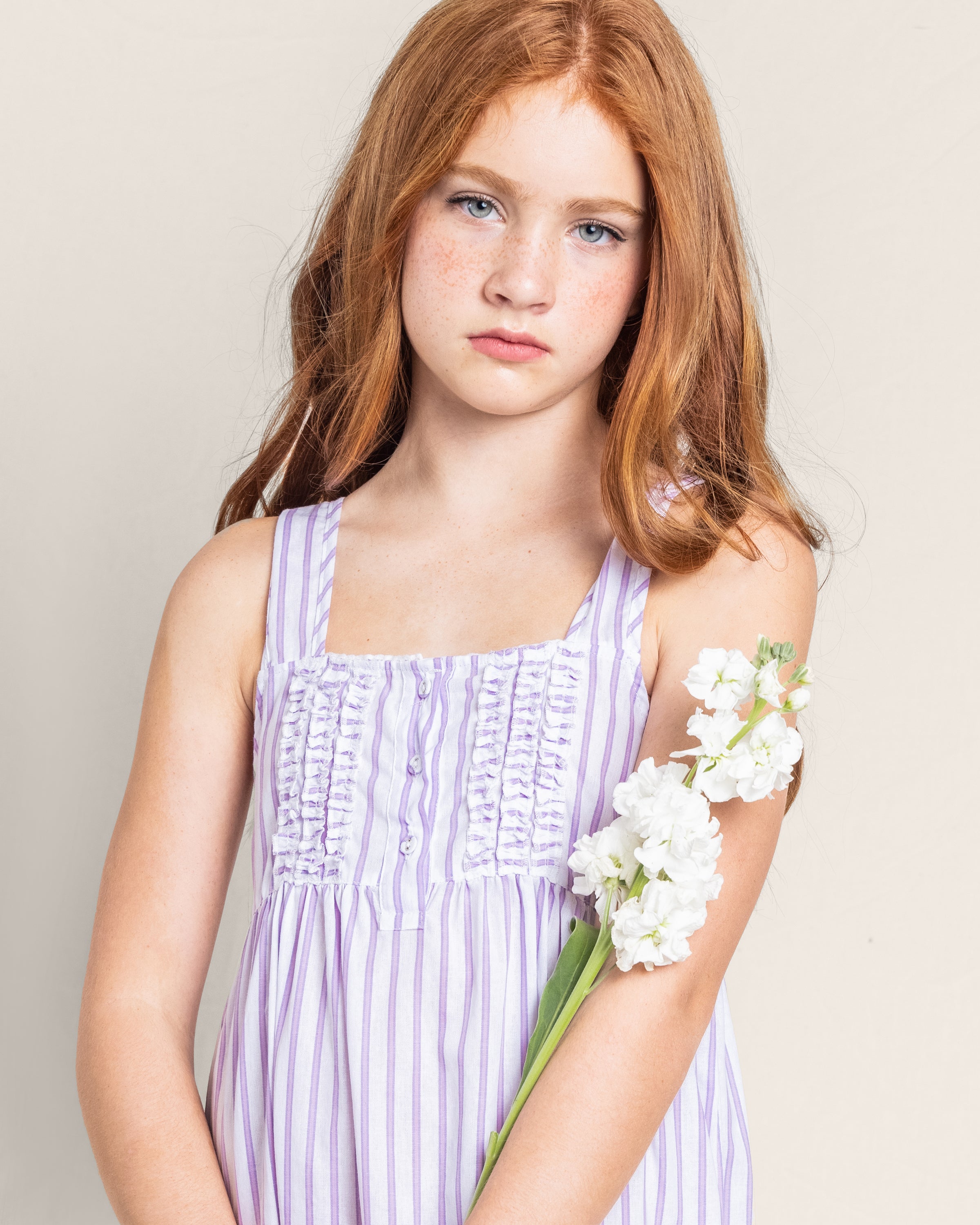 A girl with long red hair and freckles is wearing a sleeveless Petite Plume Girls Twill Charlotte Nightgown in Lavender French Ticking. She holds white flowers and stands against a plain background, looking towards the camera with a serious expression.