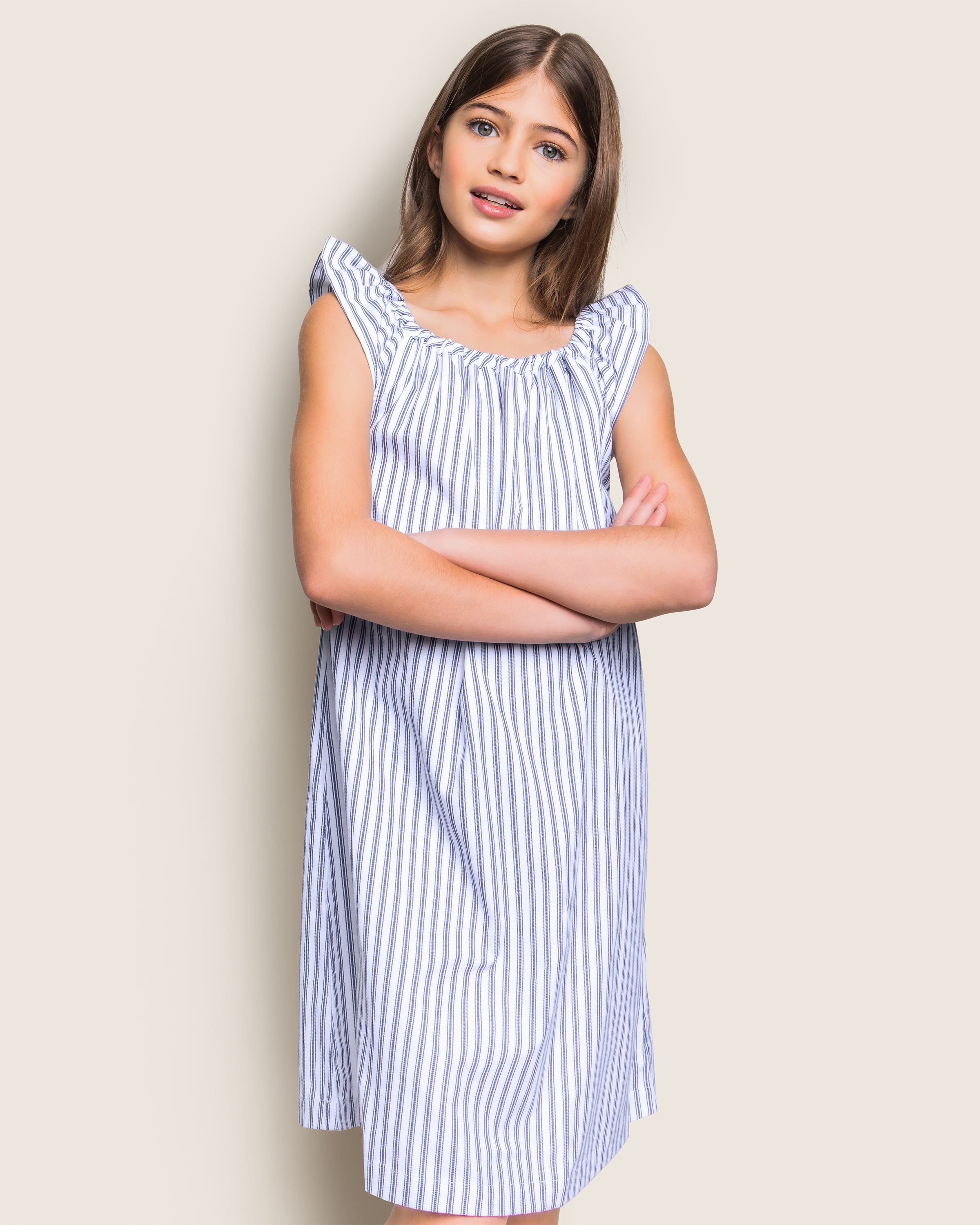 A girl with long brown hair and a neutral expression stands with arms crossed, wearing Petite Plumes Girls Twill Isabelle Nightgown in Navy French Ticking against a plain, light background.