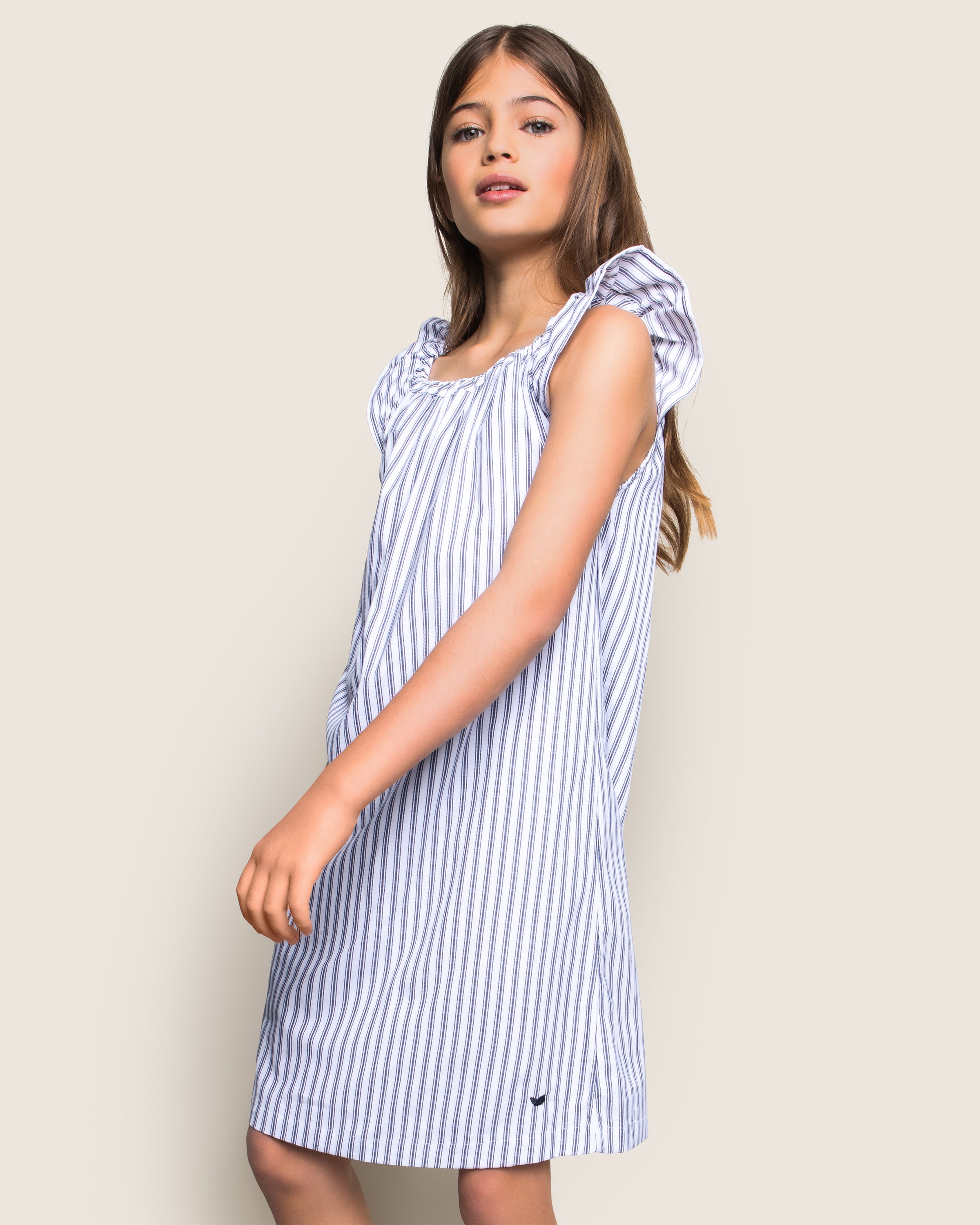 A young girl poses confidently against a neutral background in Petite Plume’s Girls Twill Isabelle Nightgown in Navy French Ticking, featuring blue and white stripes with ruffled sleeves. Her long hair cascades elegantly over her shoulders as she gazes slightly to the side.