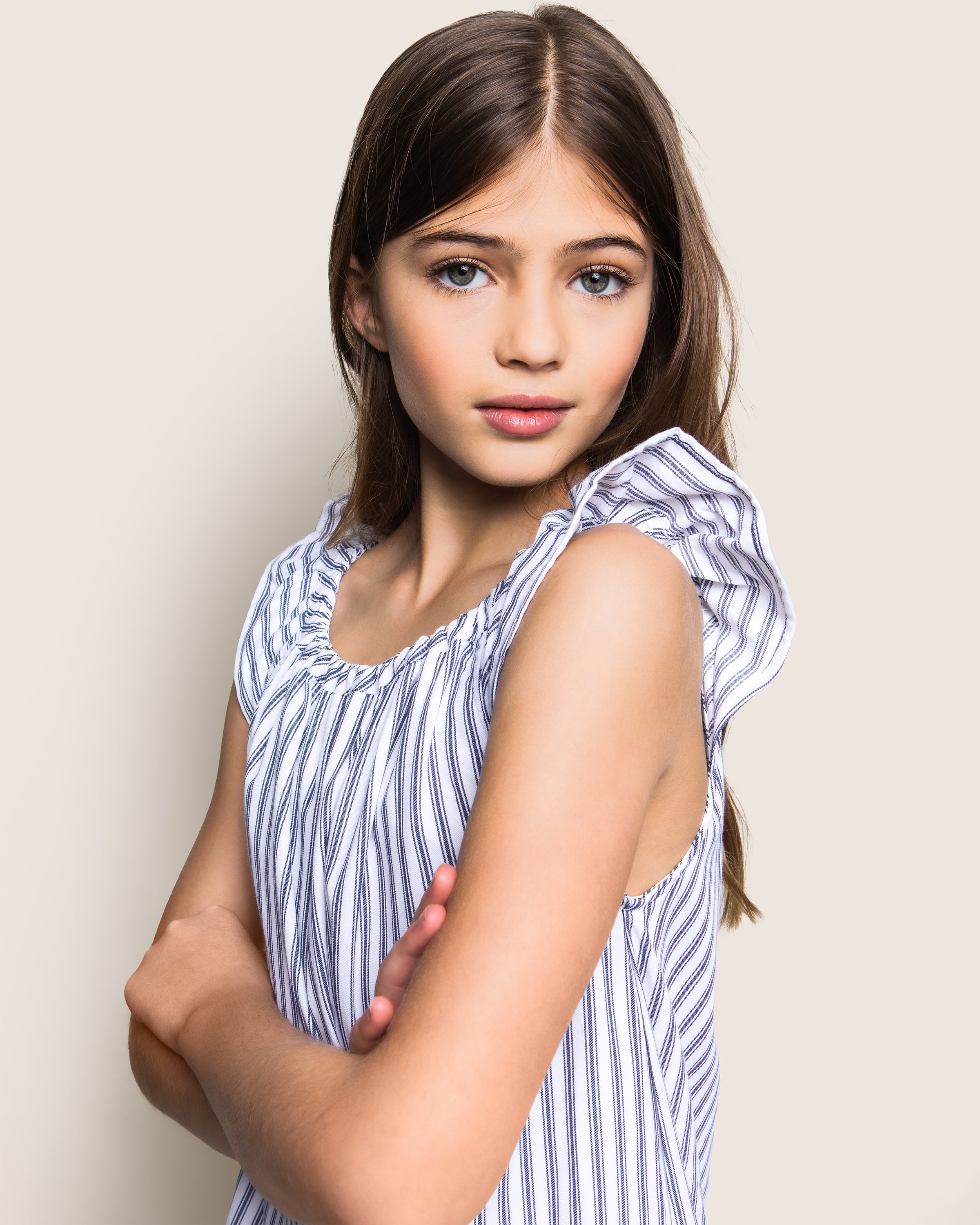 A confident young girl with long brown hair and blue eyes poses with arms crossed, wearing a Petite Plume Girls Twill Isabelle Nightgown in Navy French Ticking, set against a neutral background.