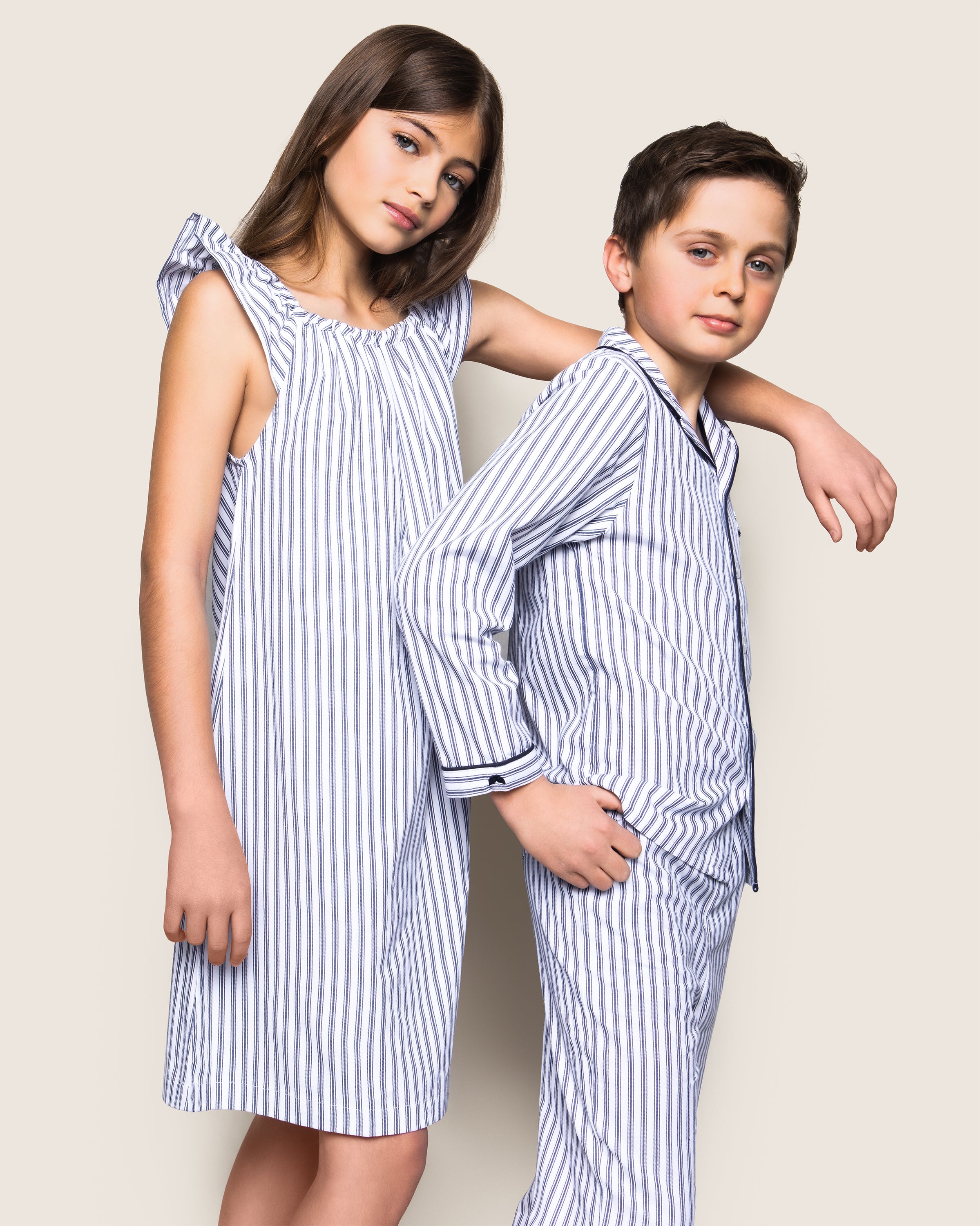 Two children pose against a plain background in matching blue and white striped pajamas by Petite Plume. The girl wears a navy French Ticking Isabelle nightgown, and the boy has a long-sleeve top with pants, both looking relaxed as he leans on her shoulder.