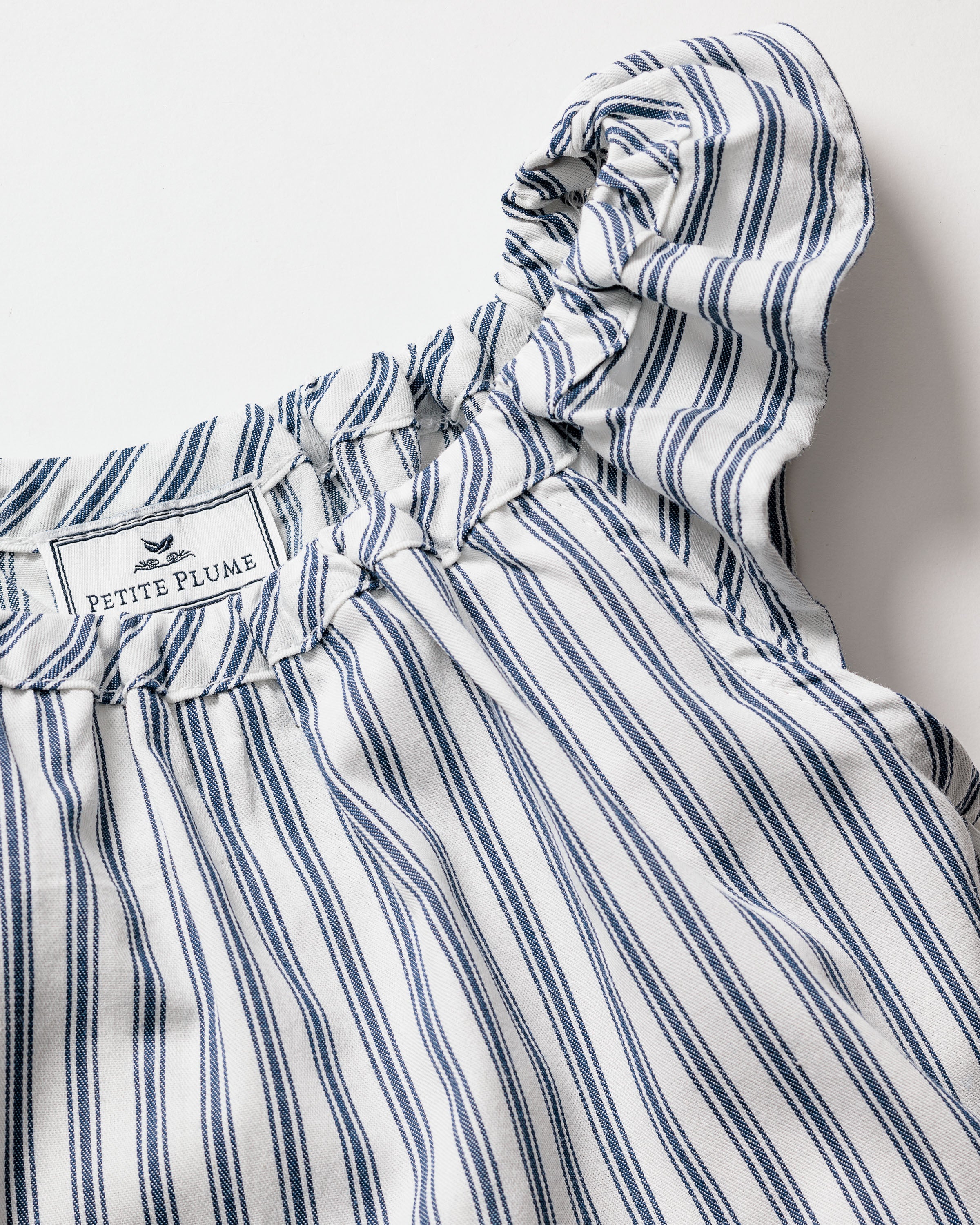 A close-up of the Petite Plume Girls Twill Isabelle Nightgown in Navy French Ticking showcases a blue and white striped design with ruffled neckline detailing. The yarn-dyed cotton fabric offers a soft, textured look for comfort and style in sleepwear.
