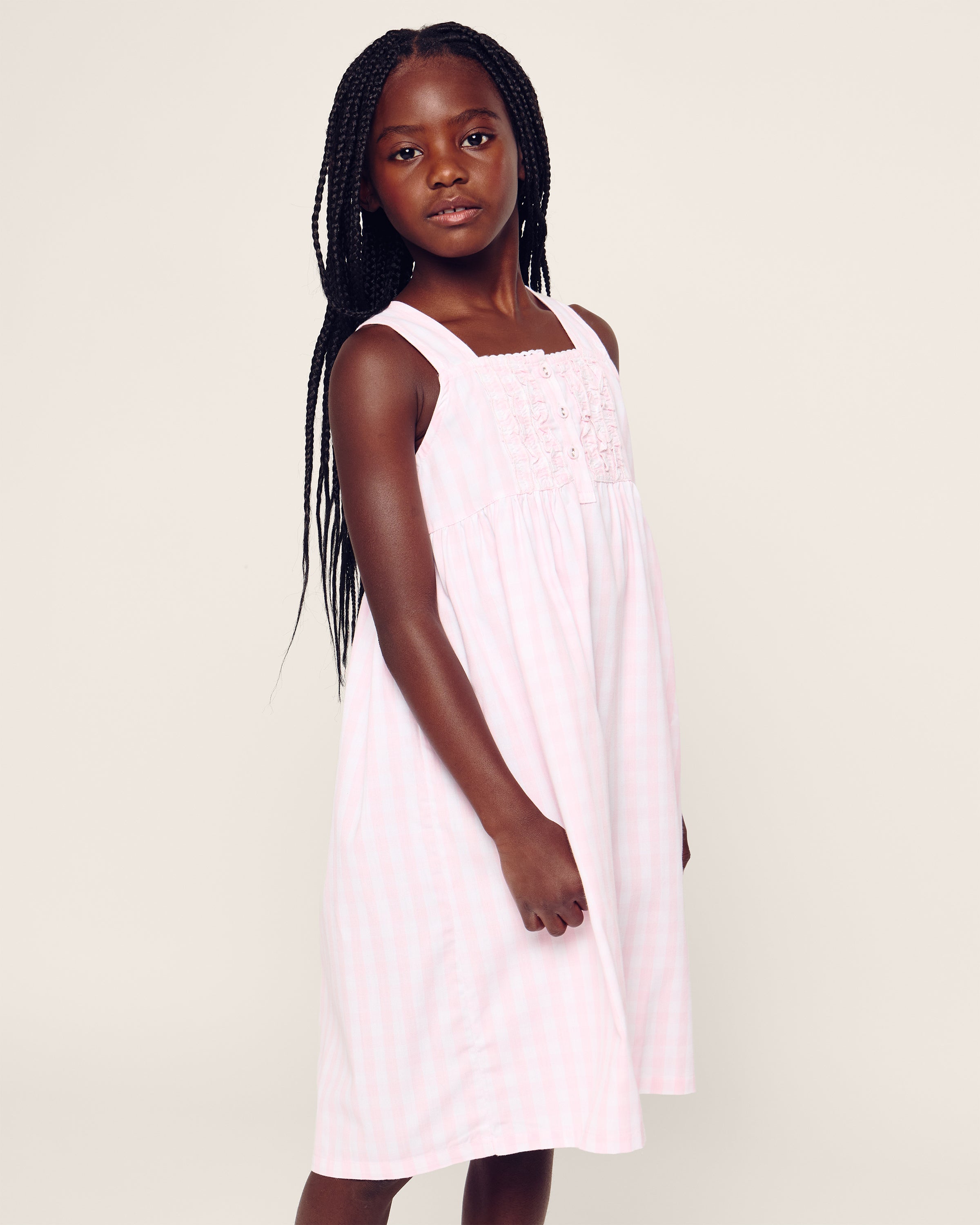 A young girl with long braided hair wears Petite Plumes Twill Charlotte Nightgown in Pink Gingham. She stands against a plain background, gazing at the camera with a neutral expression.