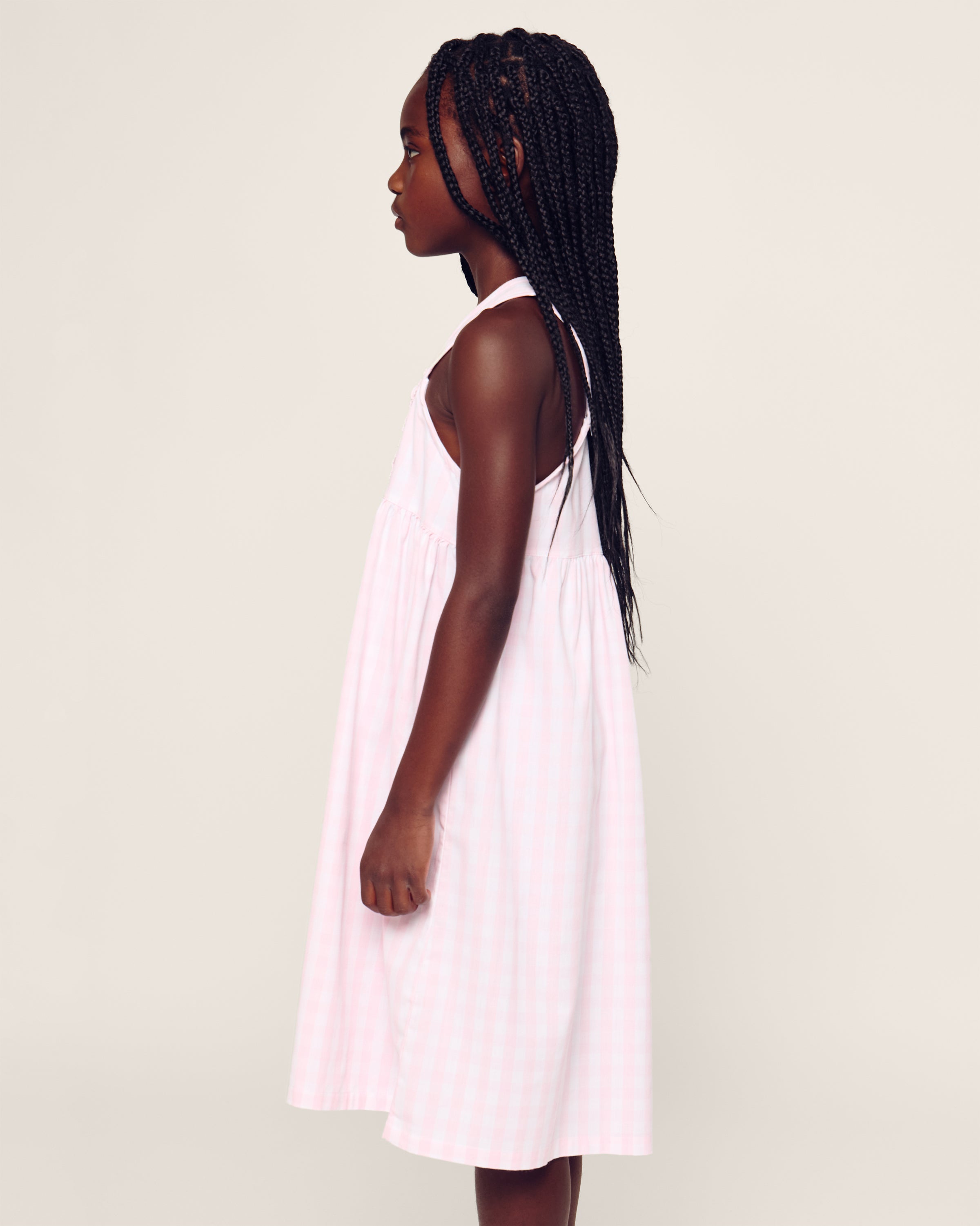 A young girl with long braided hair wears a Petite Plume Girl’s Twill Charlotte Nightgown in Pink Gingham, standing in profile against a plain light background, facing left.