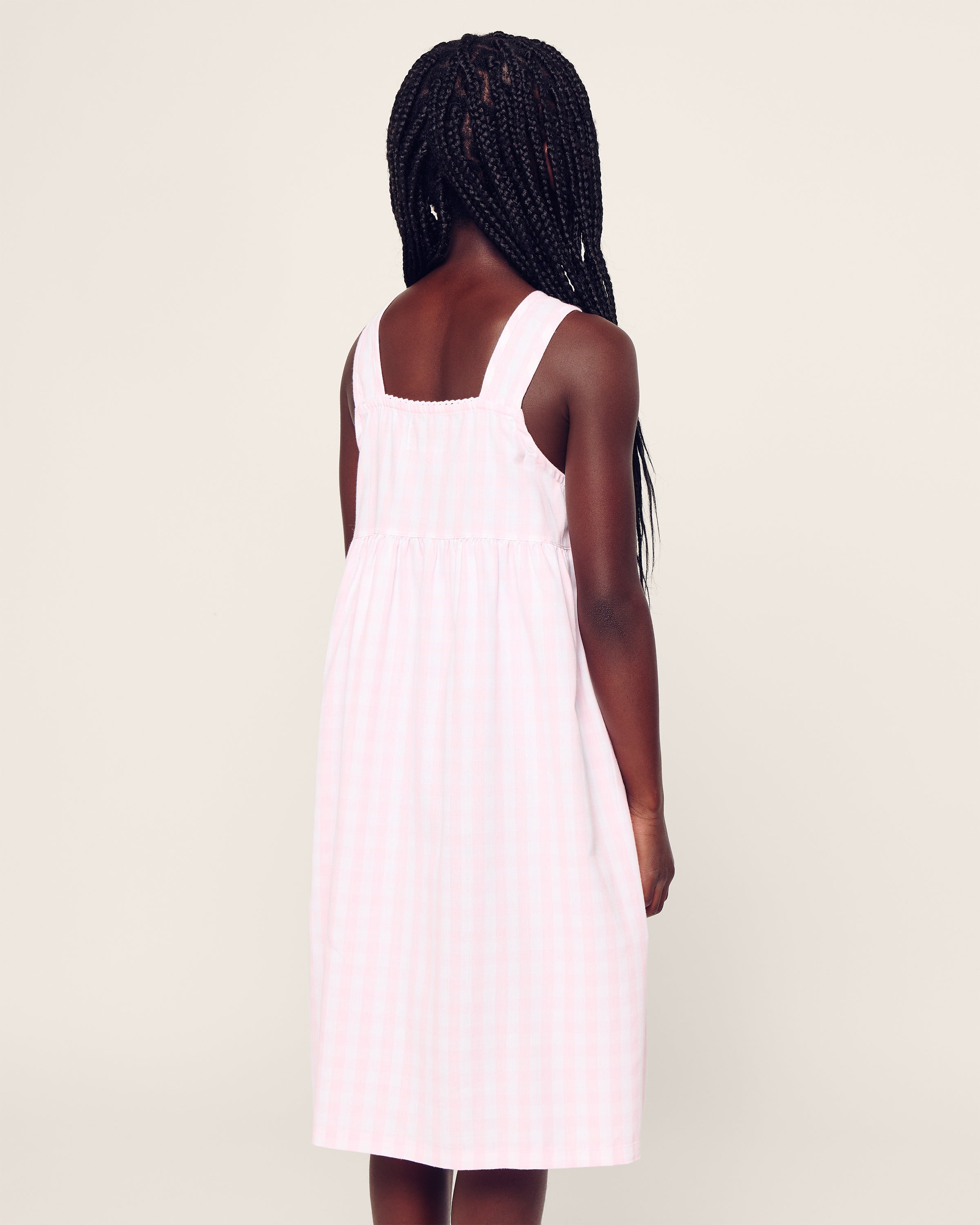 A young girl with long braided hair stands relaxed, dressed in Petite Plumes sleeveless Girls Twill Charlotte Nightgown in Pink Gingham. Crafted from soft cotton sleepwear fabric, it offers comfort and peace as she faces a plain background.