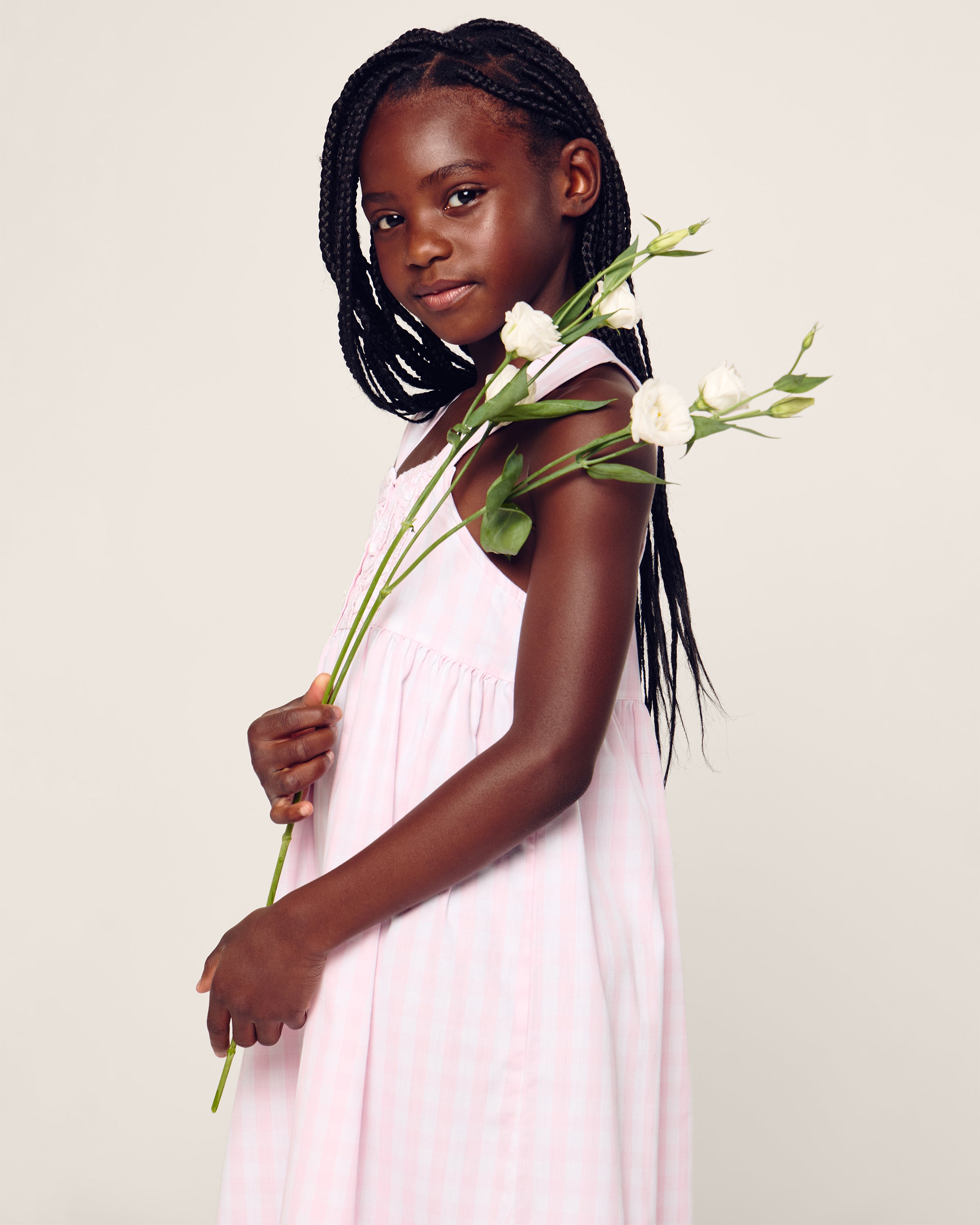 A young girl with braided hair wears the Petite Plume Girls Twill Charlotte Nightgown in Pink Gingham. She holds white flowers, stands against a plain background, and looks at the camera with a gentle expression.