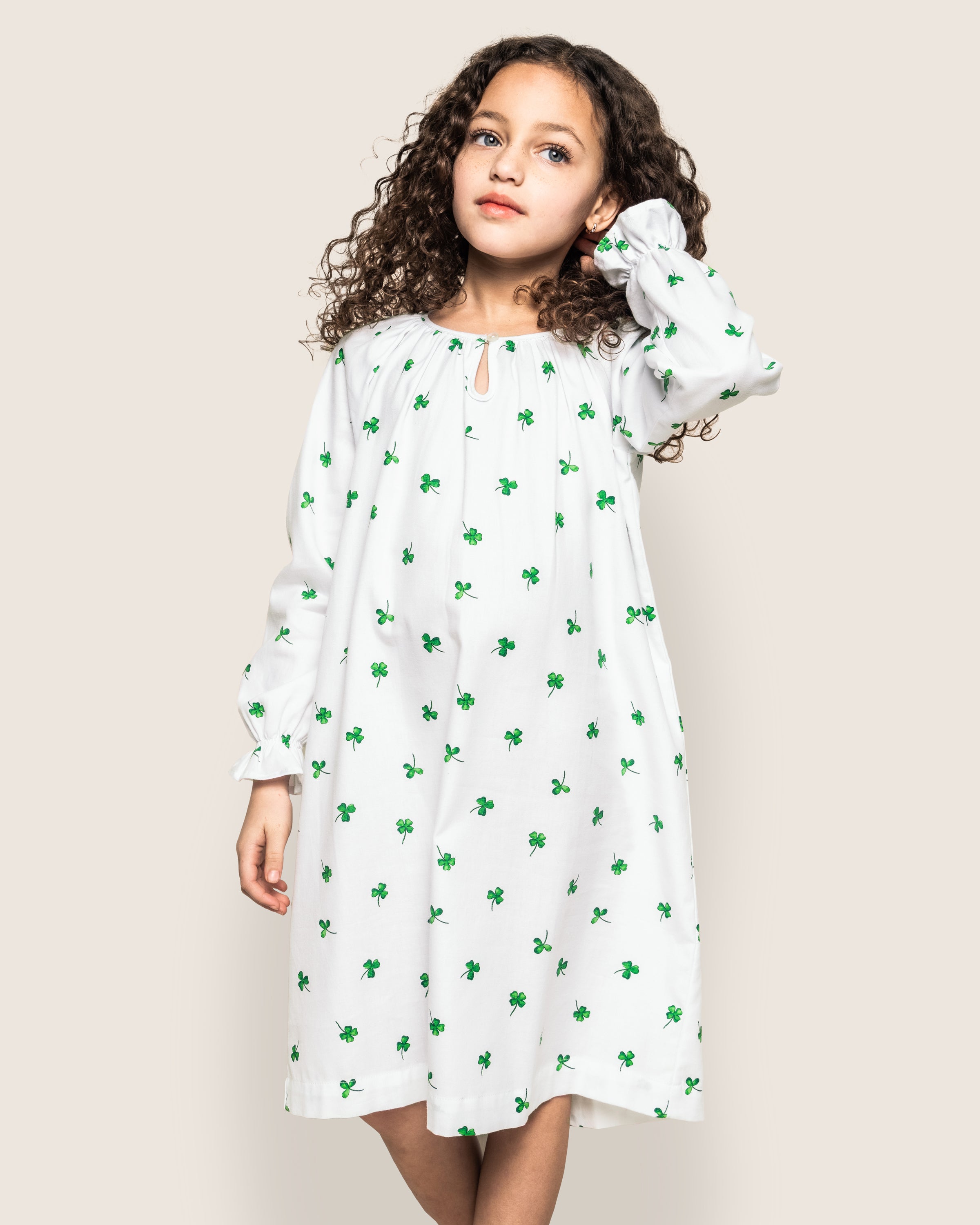 In a Petite Plume Girls Twill Delphine Nightgown from the Shamrocks Collection, the young girl wears a white dress with green plant patterns. She stands against a plain background, one hand on her curly hair and the other at her side.