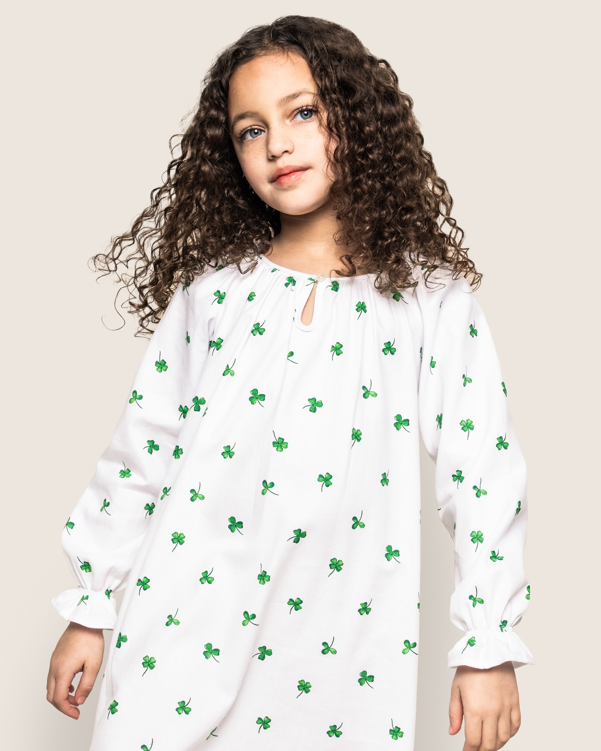 A young girl with long, curly hair wears Petite Plumes Girls Twill Delphine Nightgown in Shamrocks as she stands against a plain, light-colored background, gazing slightly to the side.