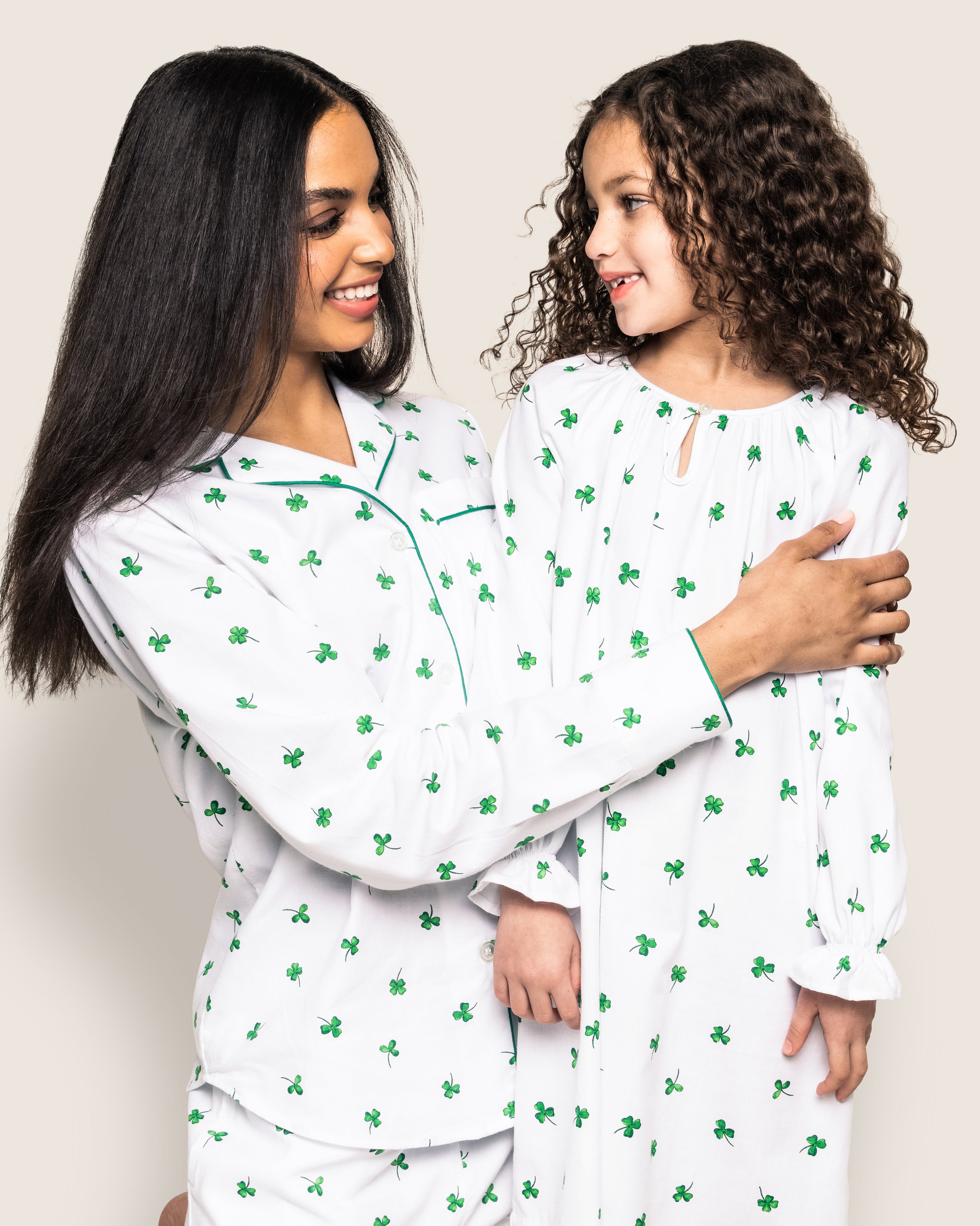 A woman and child, both with curly hair, warmly smile in matching outfits from Petite Plumes Shamrocks Collection. The woman wears a luxurious pajama set, while the child dons the Girls Twill Delphine Nightgown in Shamrocks.
