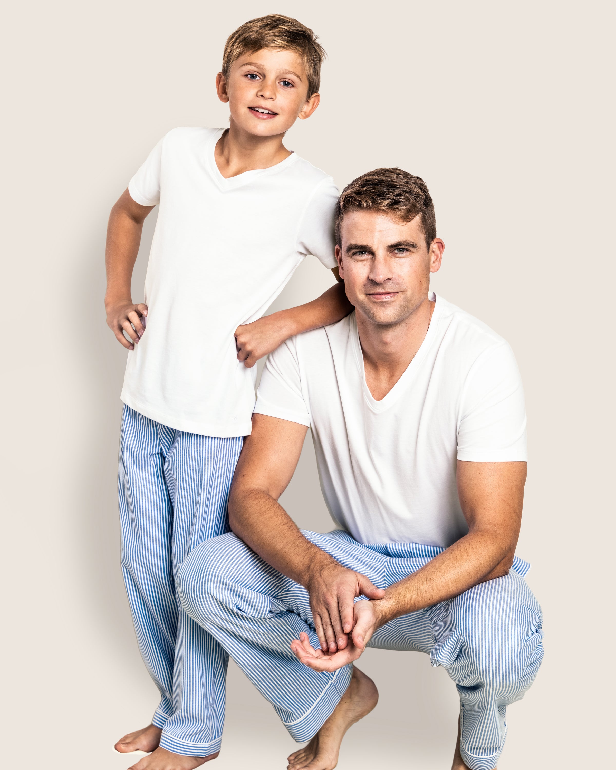 A boy and a man, both barefoot and smiling against a plain background, wear white t-shirts with Petite Plumes blue striped sleepwear: the boy in Kids Twill Pajama Pants in French Blue Seersucker stands while the man crouches beside him.