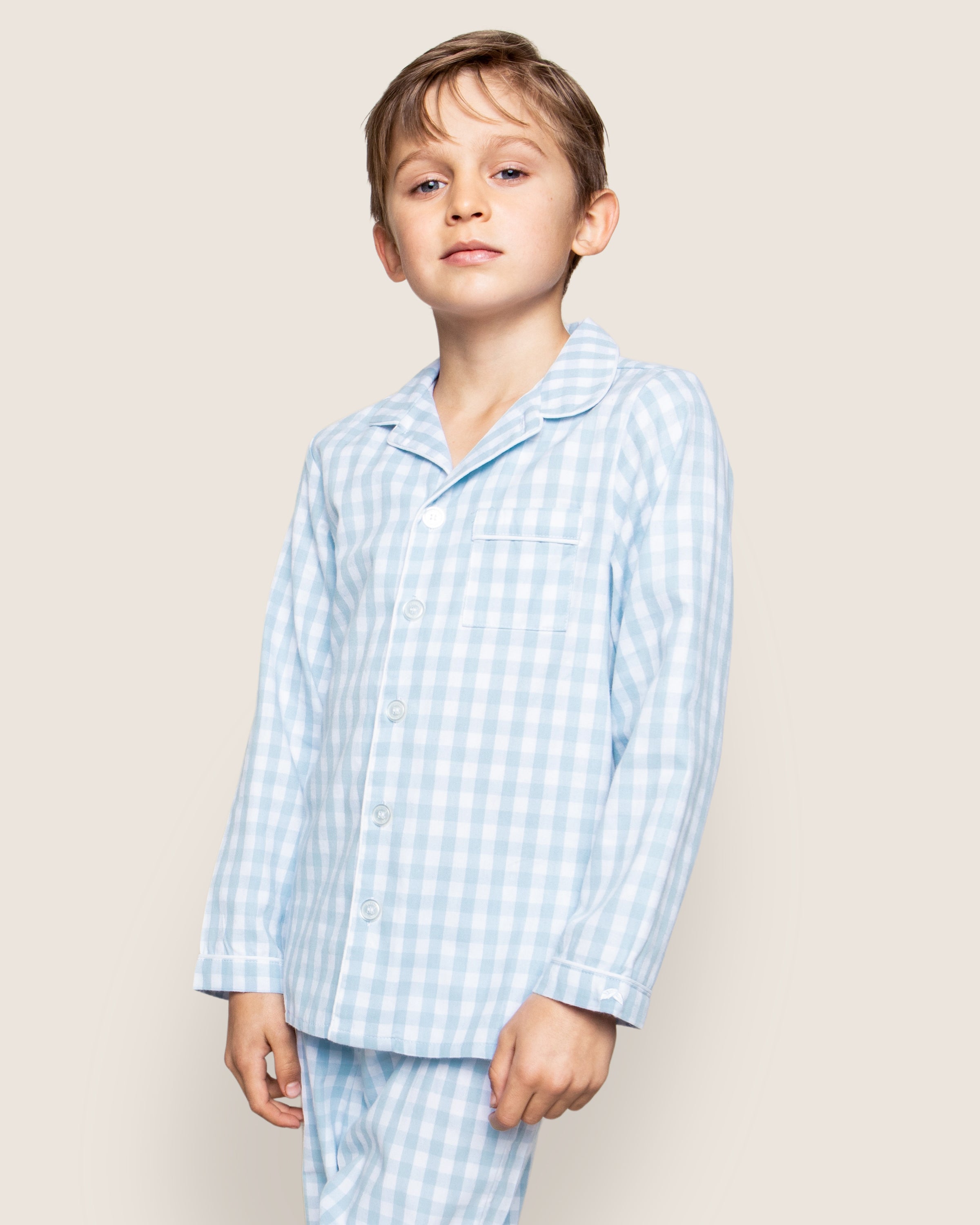 A young boy stands confidently in Petite Plumes Kids Twill Pajama Set in Light Blue Gingham. The button-up top with a collar adds charm, and its moisture-wicking twill weave ensures comfort. The plain off-white background highlights the simplicity of his chemical-free sleepwear.