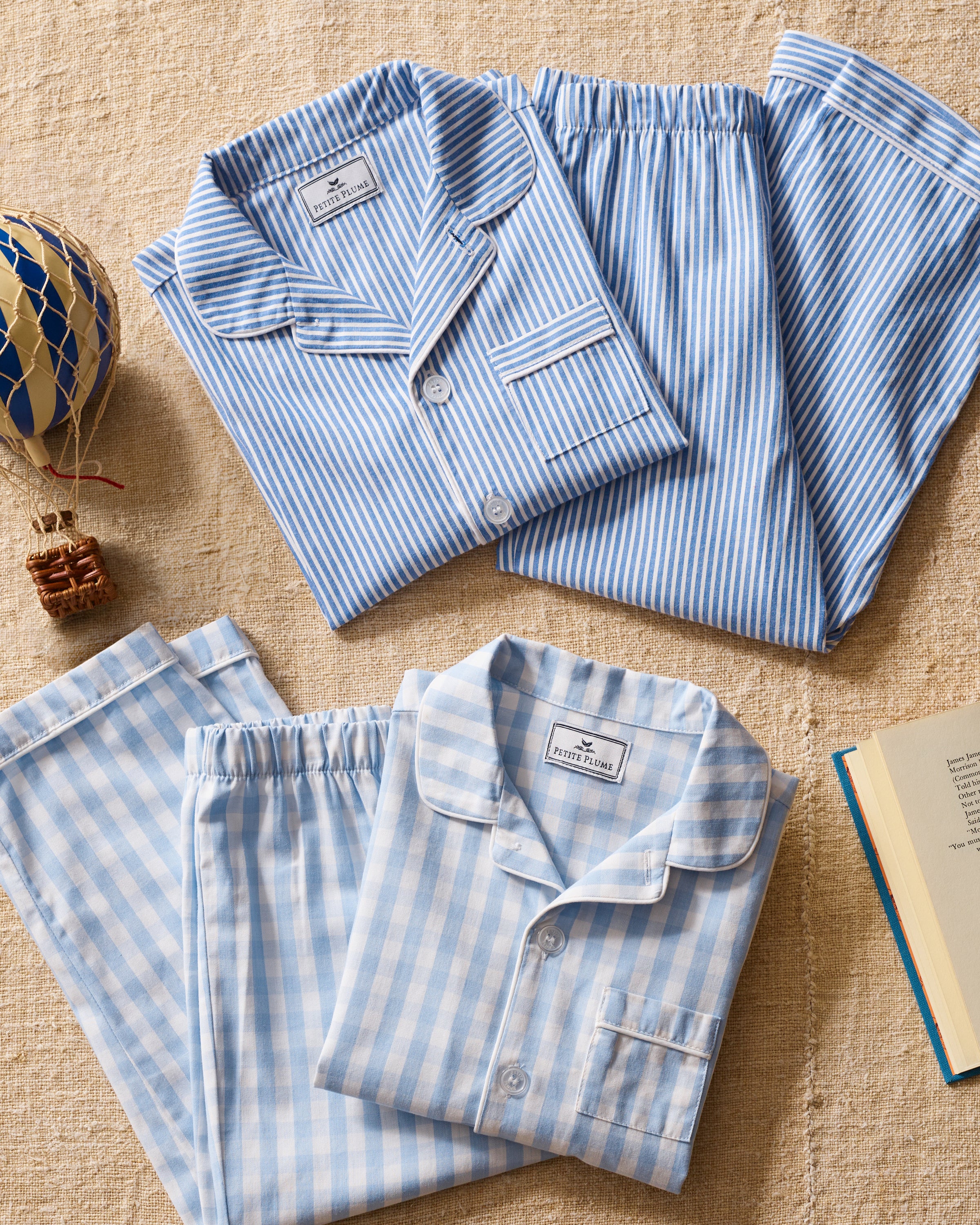 Two sets of Petite Plume classic pajamas rest on a textured beige surface. One is light blue with white stripes and the other is gingham. A small hot air balloon model and an open book accompany the flame-retardant Kids Twill Pajama Set in Light Blue Gingham for decorative accents.