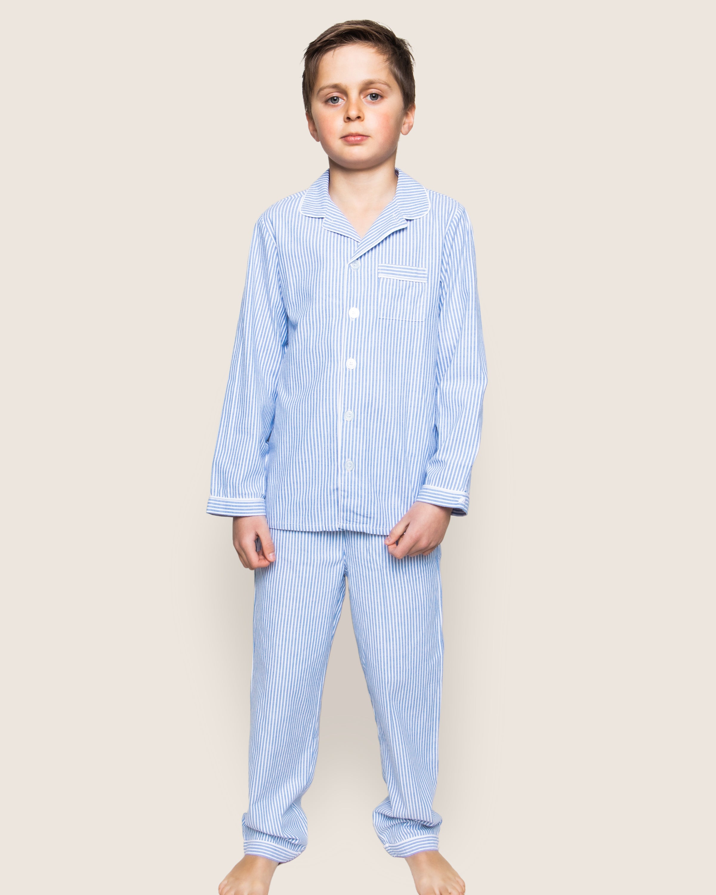 A young boy stands barefoot against a light background, wearing the refined Petite Plume Kids Twill Pajama Set in French Blue Seersucker. His collared top and long sleeves complete the luxurious look as his neutral expression complements his relaxed stance with hands by his sides.