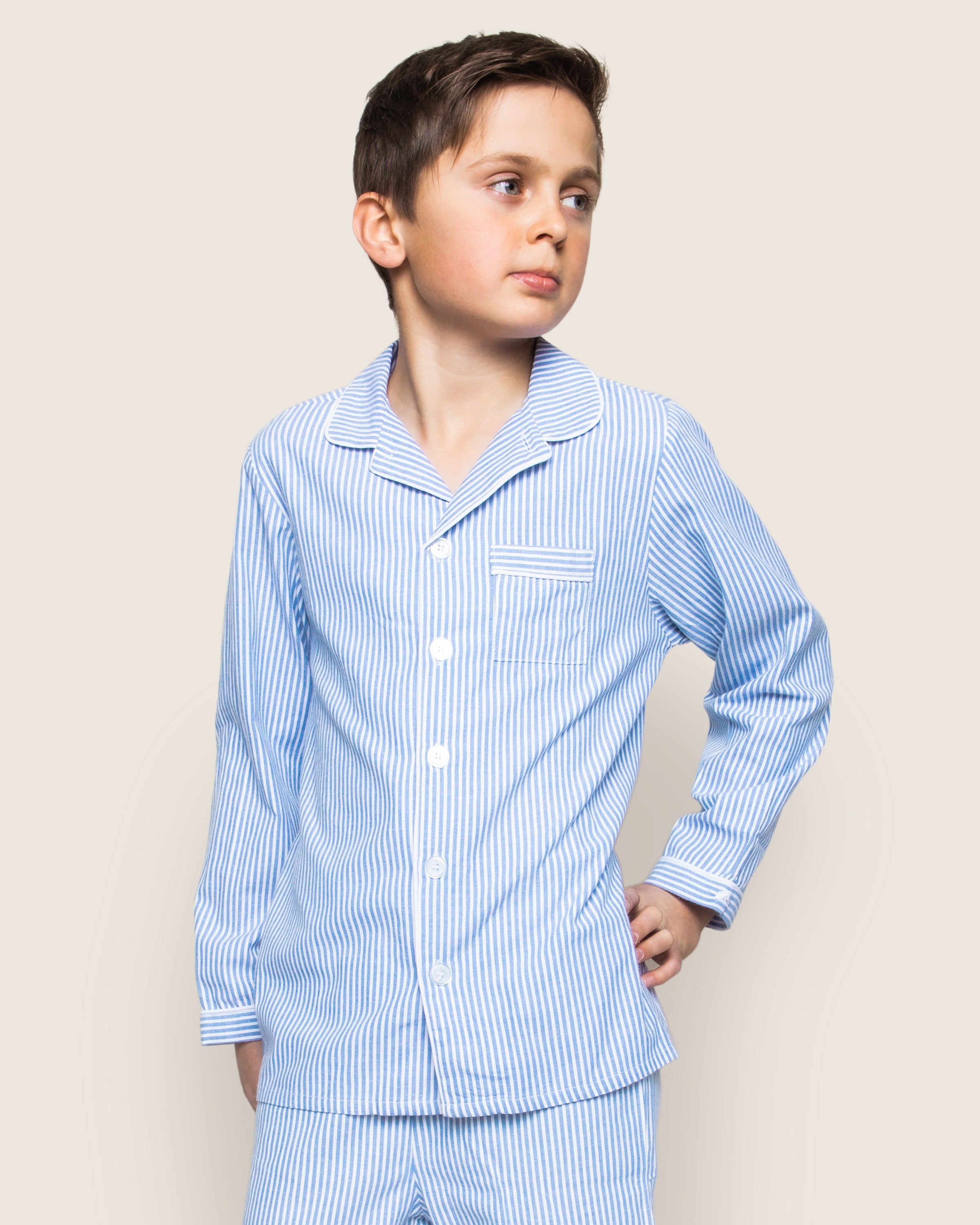 A boy in Petite Plumes Kids Twill Pajama Set in French Blue Seersucker stands against a plain background. The set includes a button-down front and chest pocket. With one hand on his hip, he gazes to the side with a neutral expression.