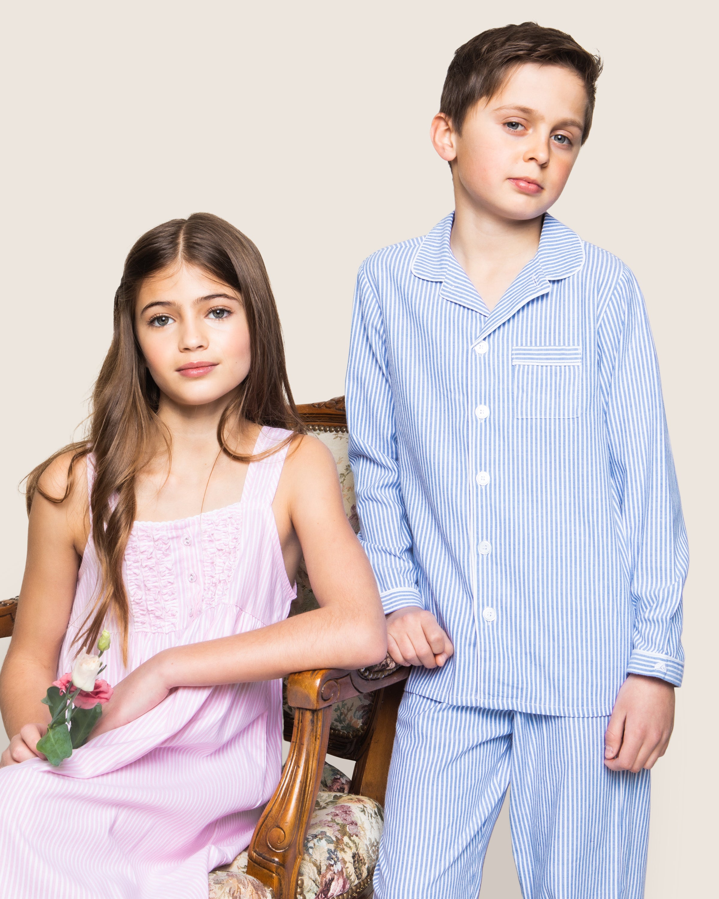 A girl in a pink nightgown sits on a floral-patterned chair with a flower, while a boy in the Petite Plume Kids Twill Pajama Set in French Blue Seersucker stands next to her against a plain background.