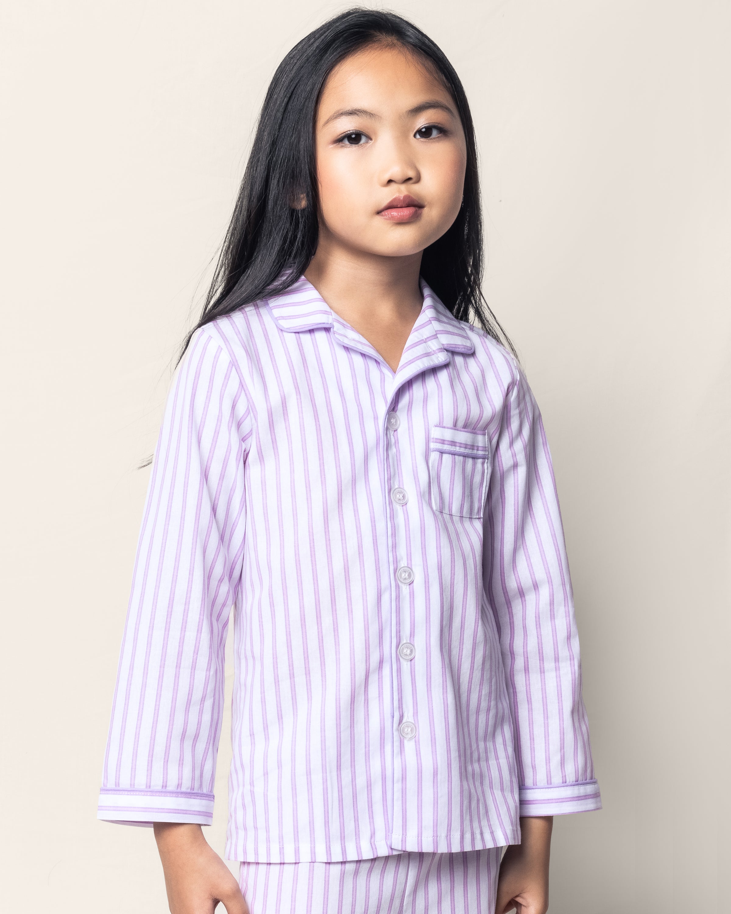 A young girl with long black hair wears the Kids Twill Pajama Set in Lavender French Ticking by Petite Plume, featuring stripes, a collar, and buttons. She stands against a plain light background, embracing the charm of luxurious sleepwear.
