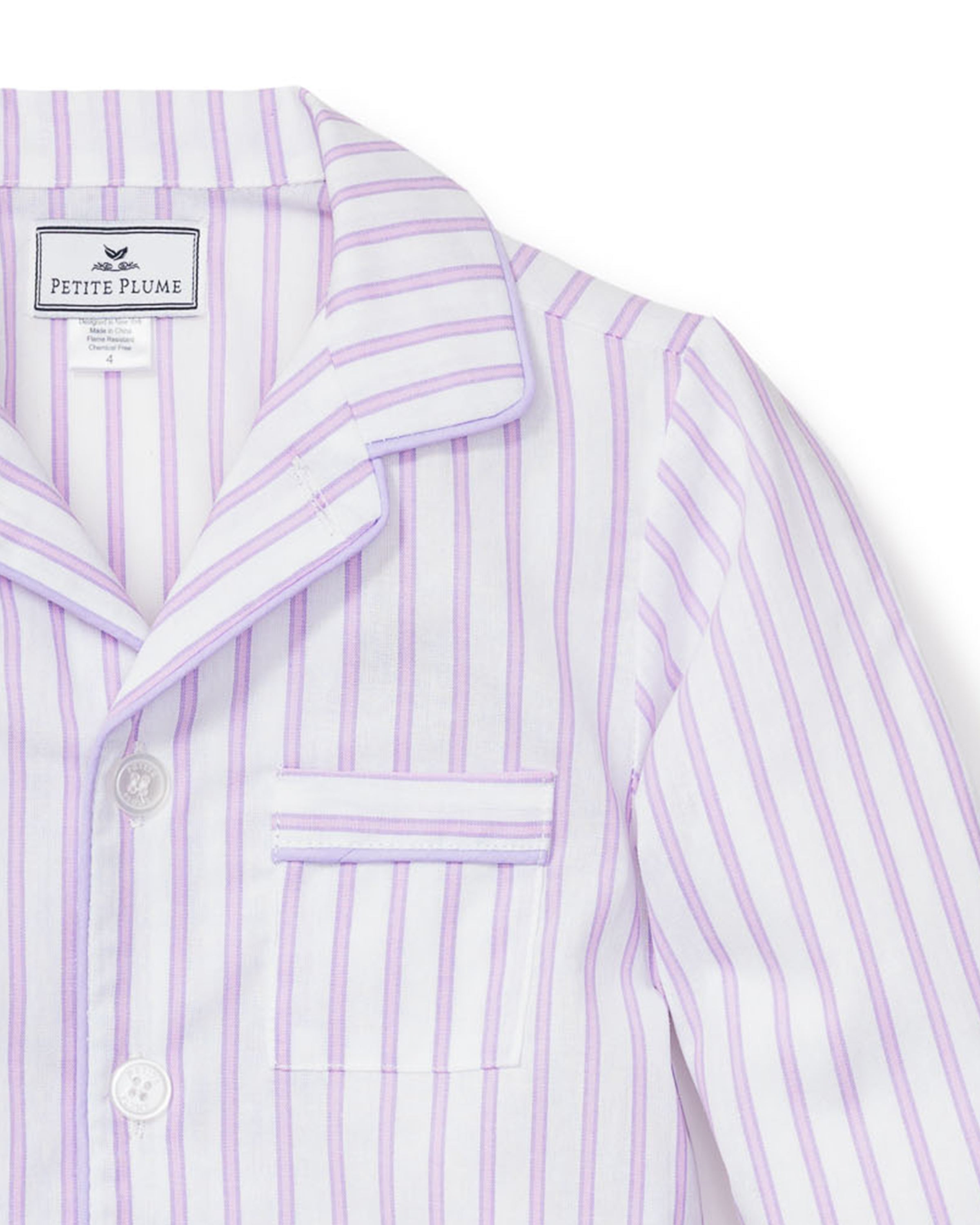 The Kids Twill Pajama Set in Lavender French Ticking by Petite Plume features a close-up of the white button-up top with stripes, a collar, pocket, and white buttons. The inside label reads Petite Plume, embodying luxury in every stitch.