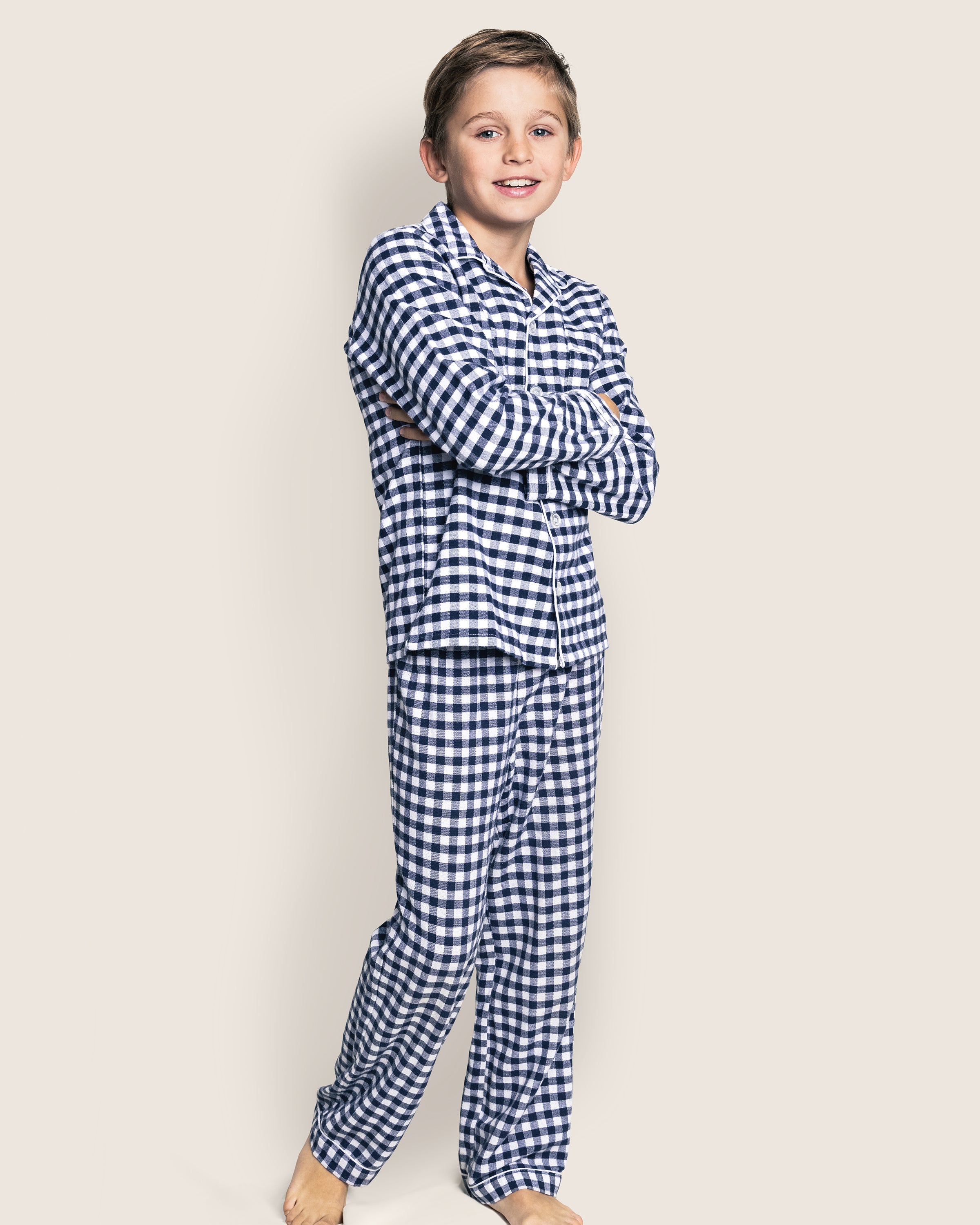 A boy stands barefoot with arms crossed, smiling in his Petite Plume Kids Twill Pajama Set in Navy Gingham against a plain light background.