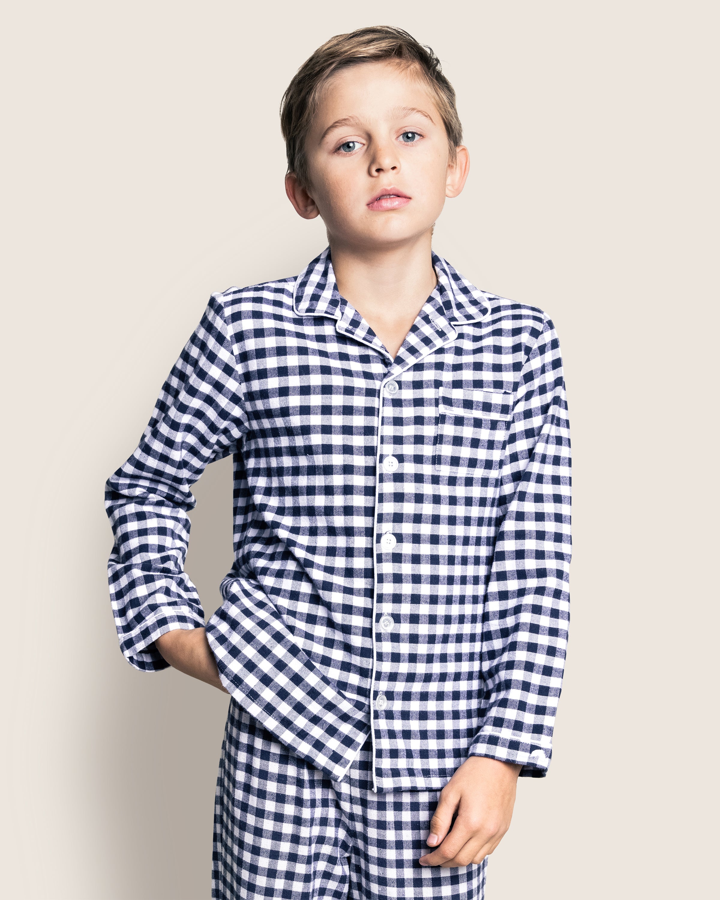 A child with short hair stands with one hand in a pocket, wearing Petite Plumes Kids Twill Pajama Set in Navy Gingham, against a plain light-colored background and looks slightly upward.