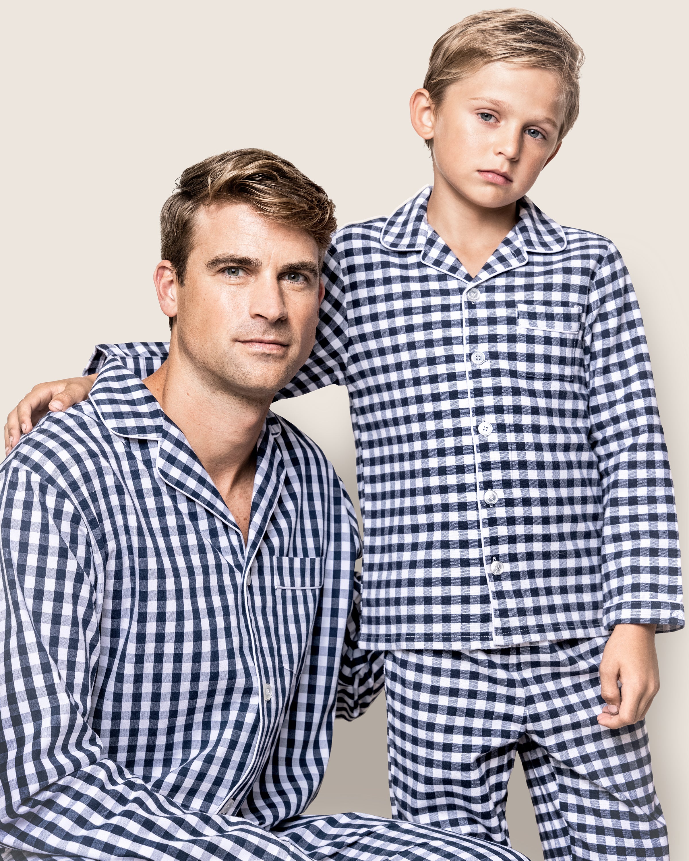 A man and boy pose seriously against a neutral background, wearing navy gingham pajama sets from Petite Plume. The man kneels with one arm around the standing boy.