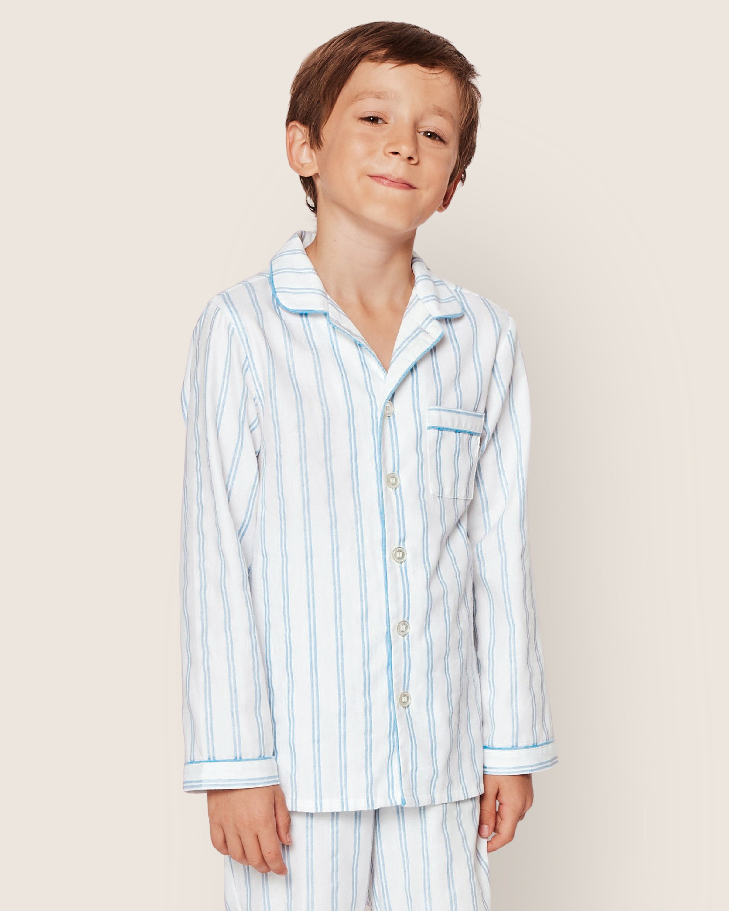 A young boy smiles softly, head tilted, wearing the Petite Plume Kids Twill Pajama Set in Periwinkle and White Stripe, standing against a plain light background.