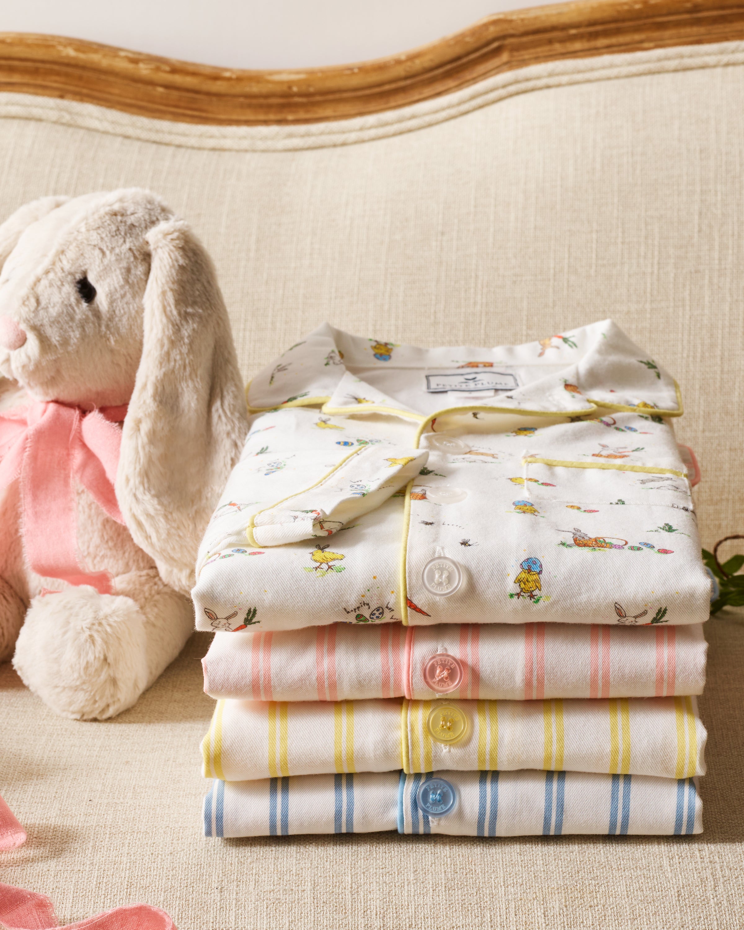 A plush white bunny with a pink ribbon sits on a beige couch next to a neatly folded Petite Plume Kids Twill Pajama Set in periwinkle and white stripe.