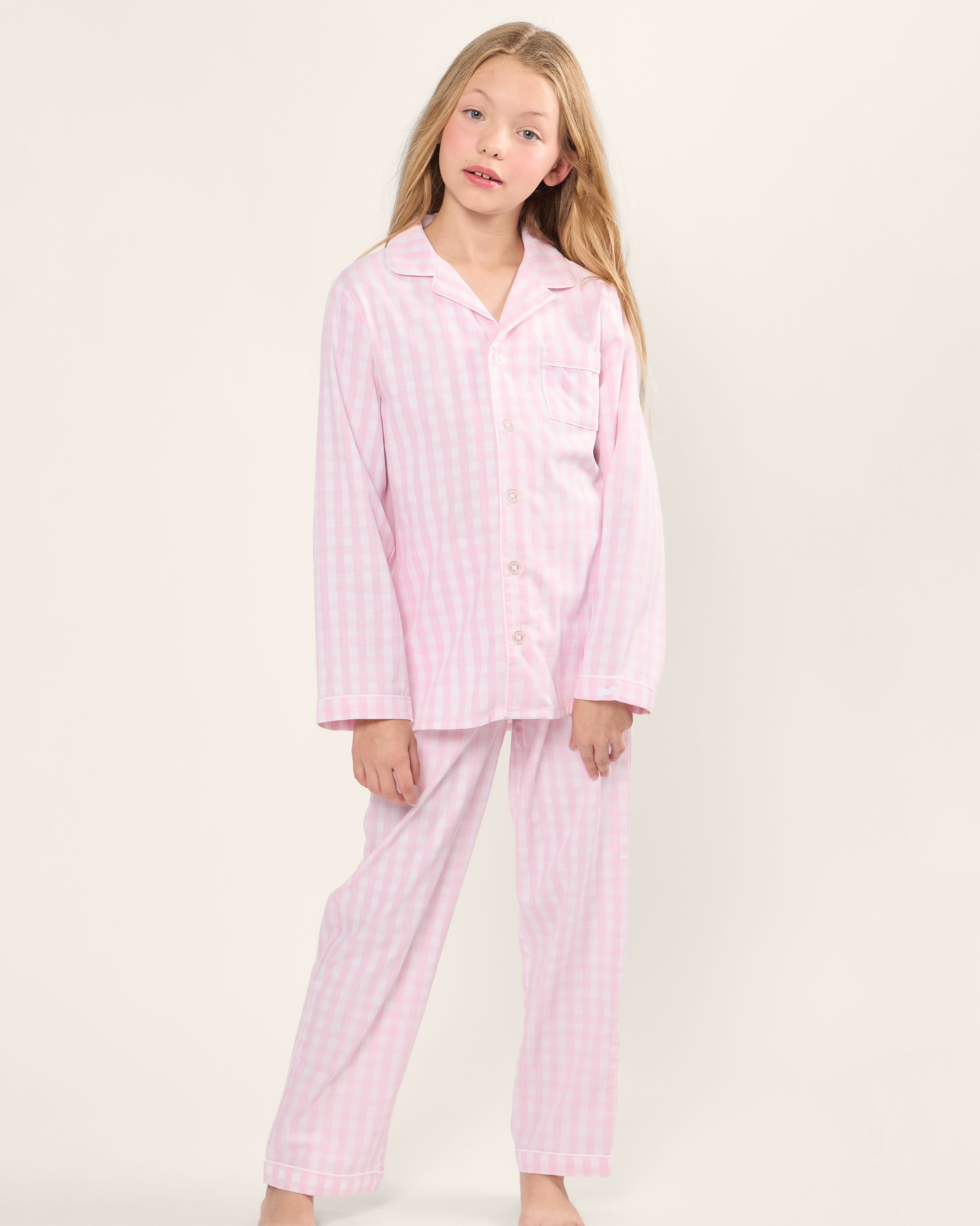 A young girl wearing Petite Plumes Kids Twill Pajama Set in Pink Gingham stands barefoot against a plain background, tilting her head slightly with a relaxed expression.